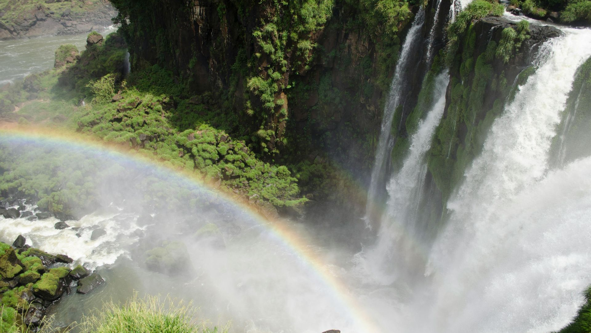 Rainbow arches over a waterfall cascading over lush green rocks.