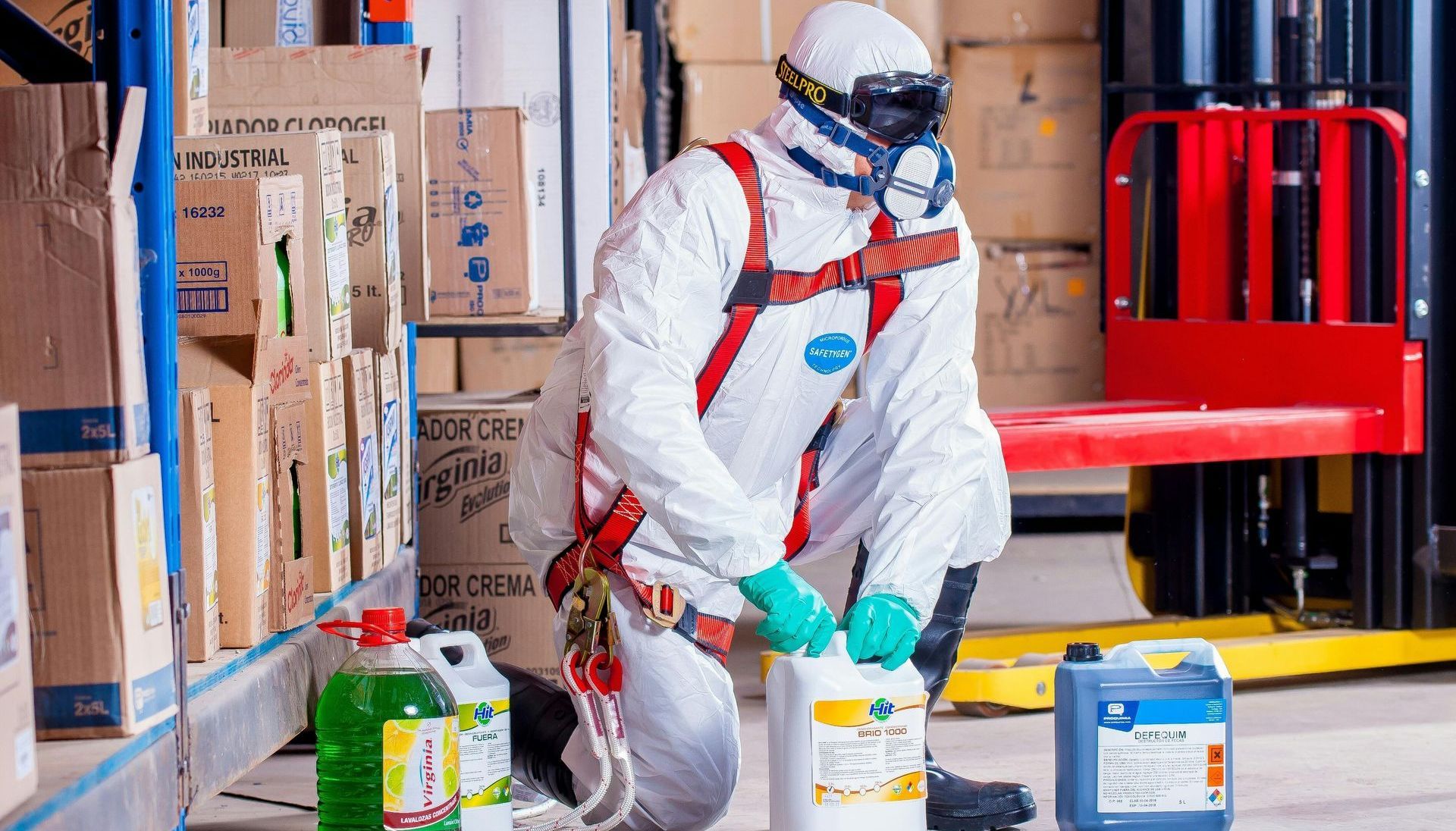 A man in a protective suit is kneeling down in a warehouse next to a forklift.