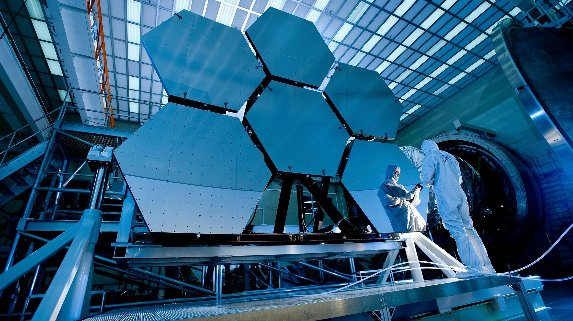 A worker in protective suit adjusts hexagonal telescope mirror segments inside a large facility.