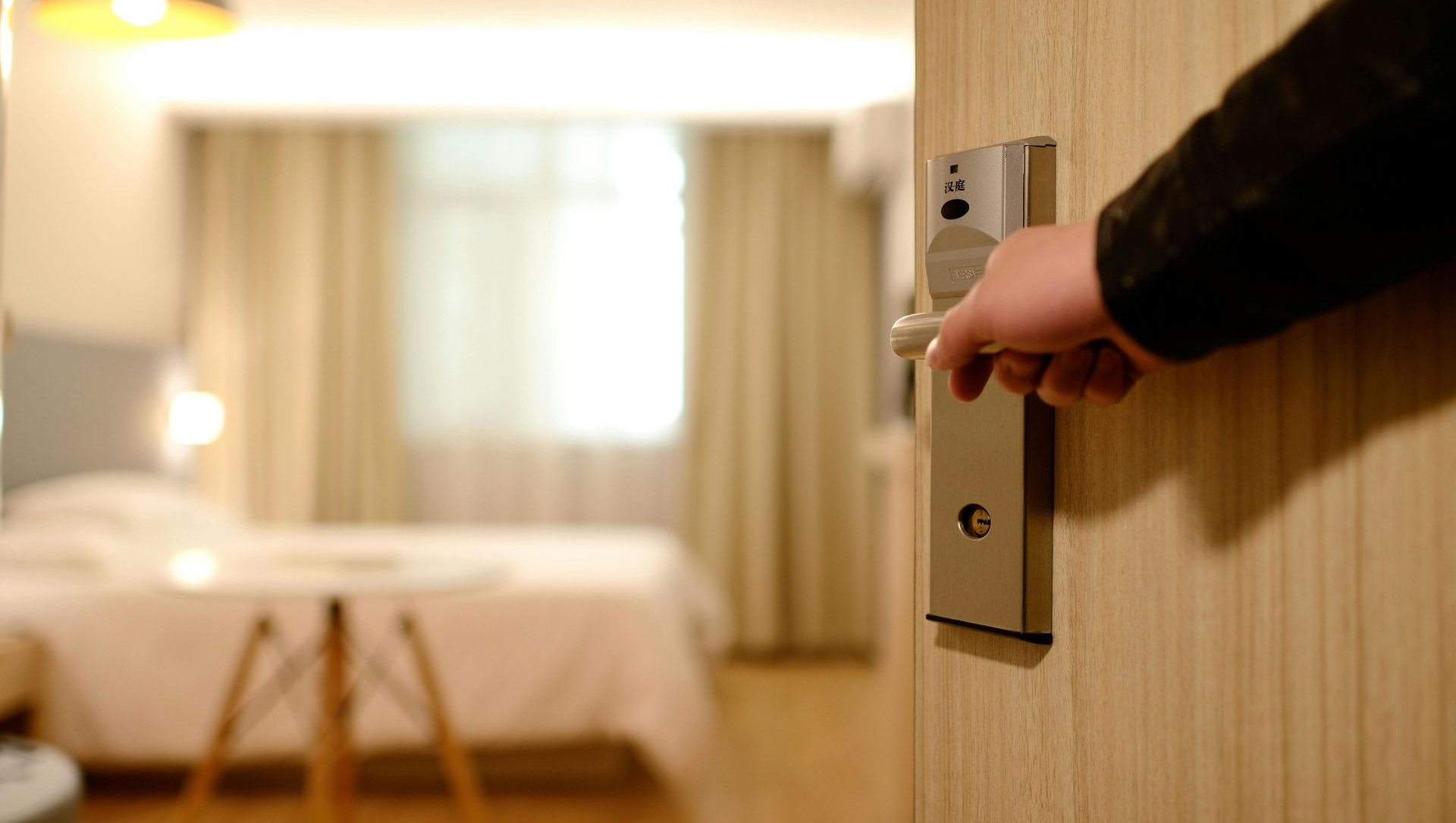 Person opening a hotel room door, blurred bedroom in the background.