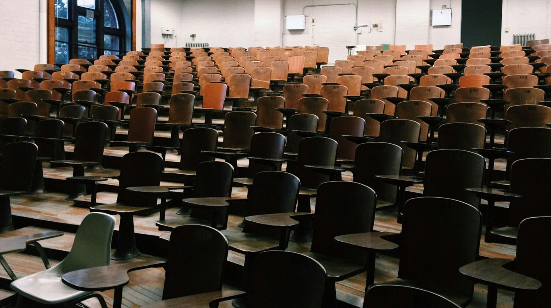 Empty lecture hall with rows of wooden desks and chairs, natural light from a large window.