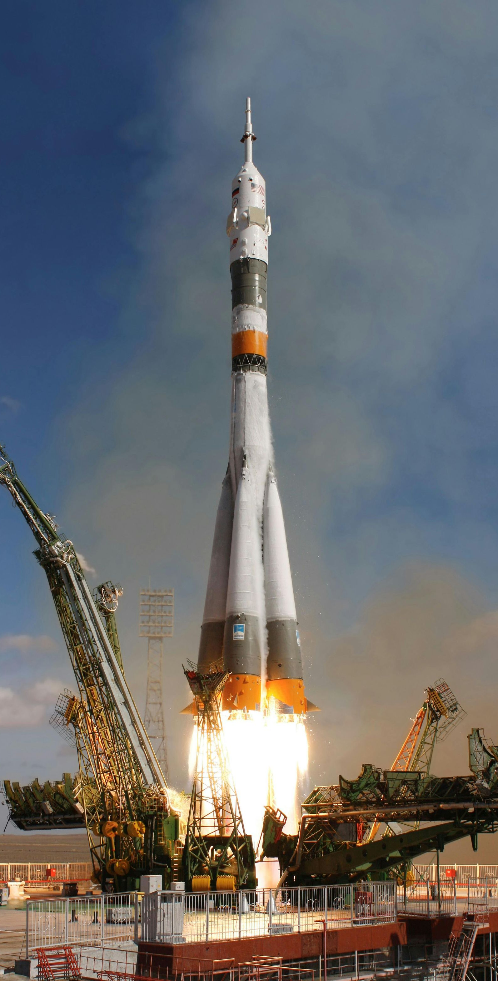 A Soyuz rocket launches from the ground, emitting flames and smoke against a blue sky.