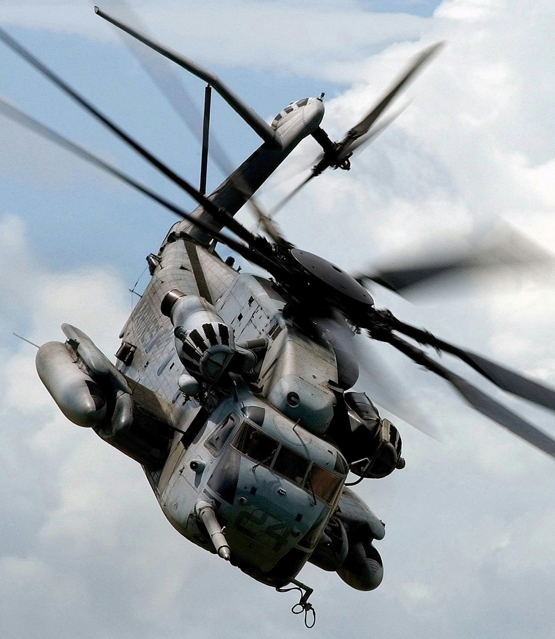 Gray military helicopter in flight, blades blurred, against a cloudy sky.