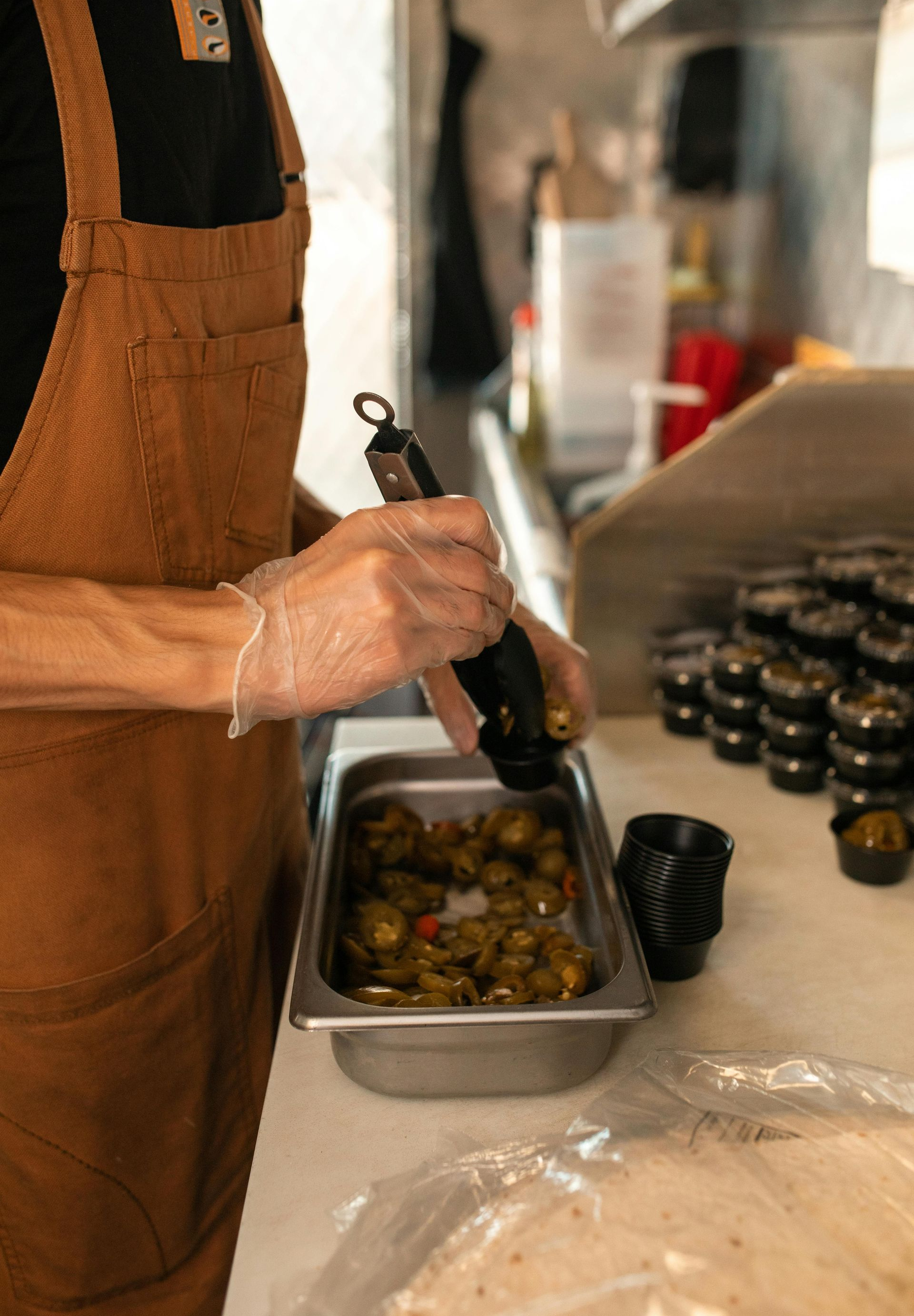 Person in apron using tongs to add food to a container in a kitchen.