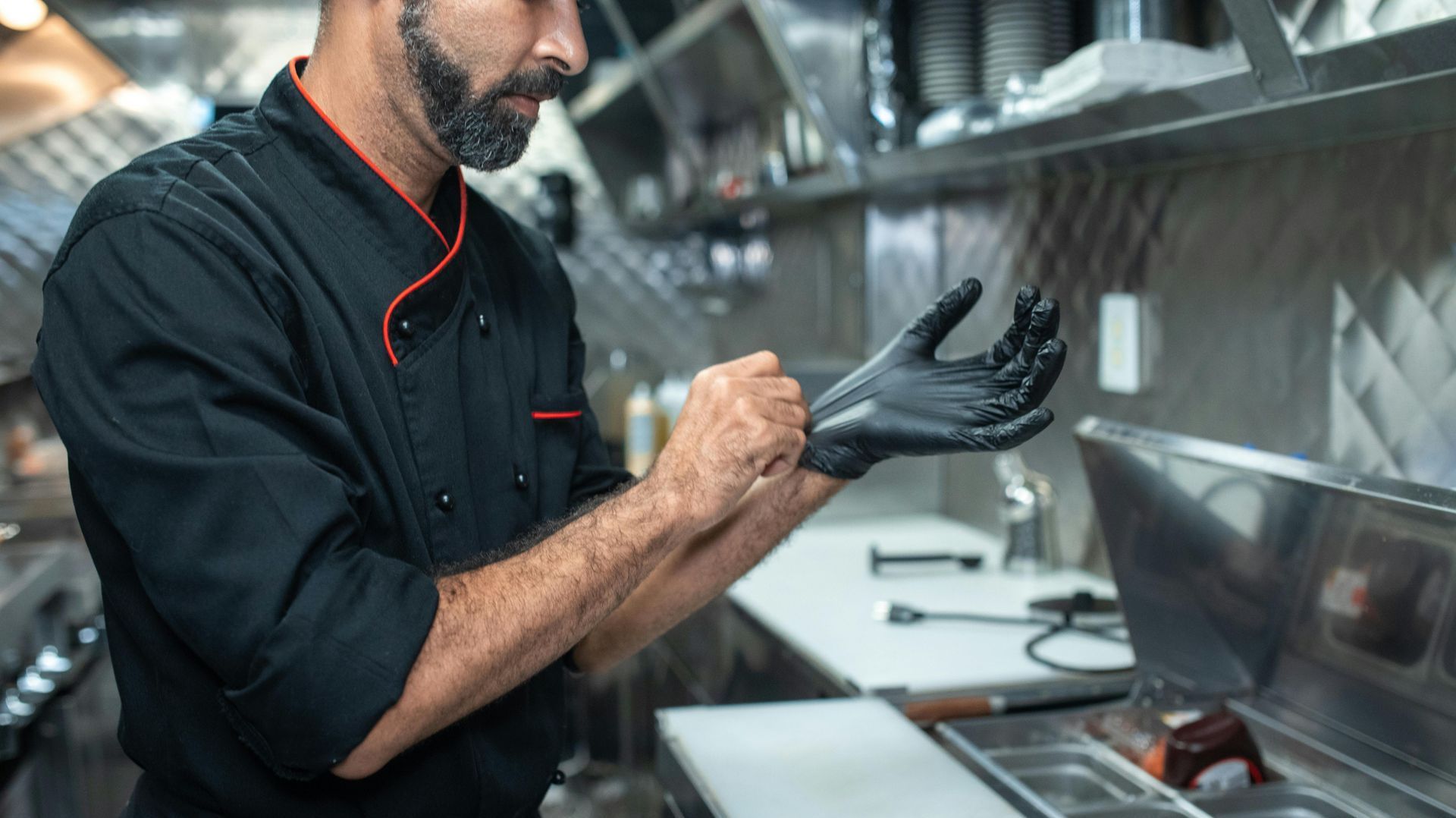 A chef is putting on a pair of black gloves in a kitchen.