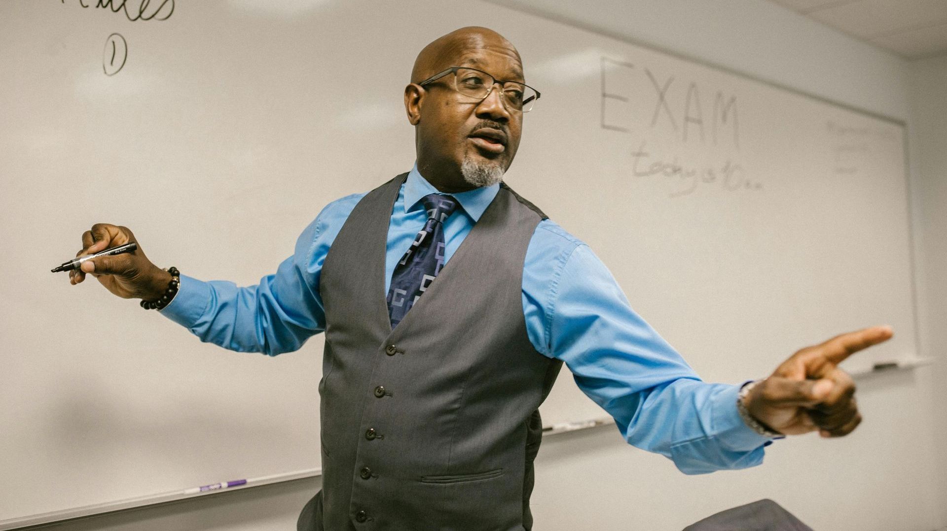 Professor in a blue shirt and gray vest gestures with a marker near a whiteboard labeled 