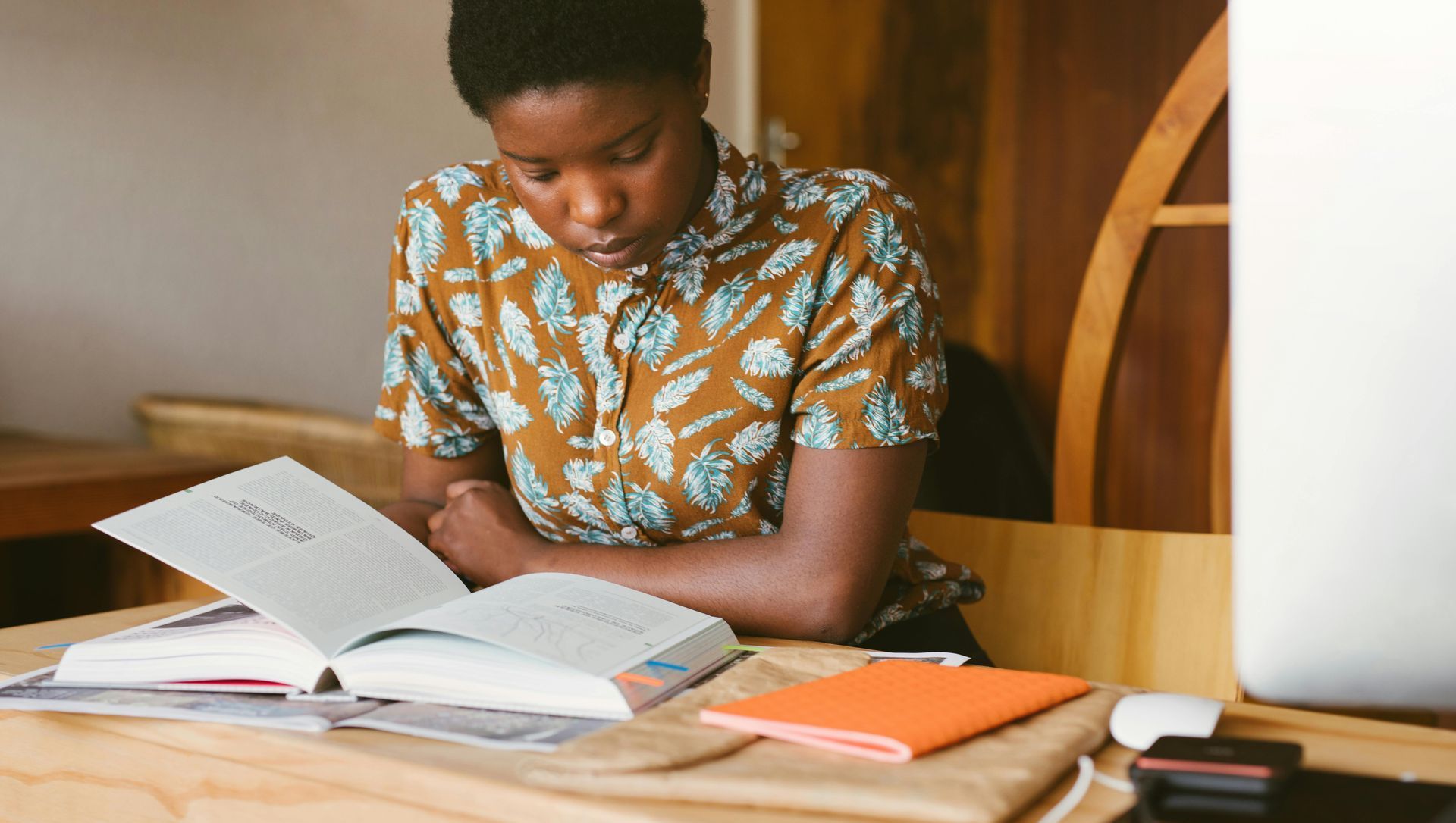 A person reads a book at a desk indoors, wearing a patterned shirt, with other books and a notebook nearby.