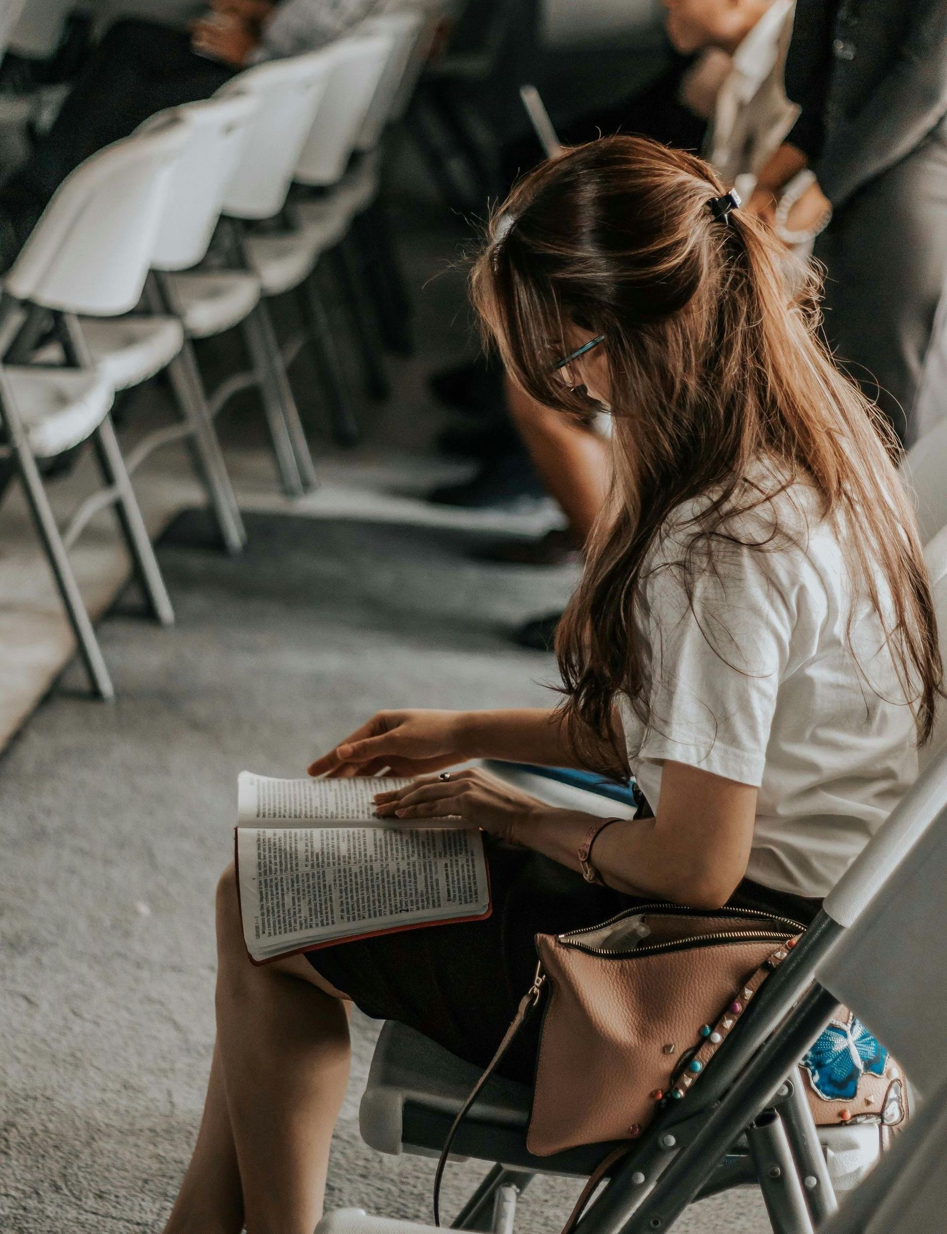 Woman with long brown hair reads a book while seated in a row of white chairs, likely at an event or gathering.