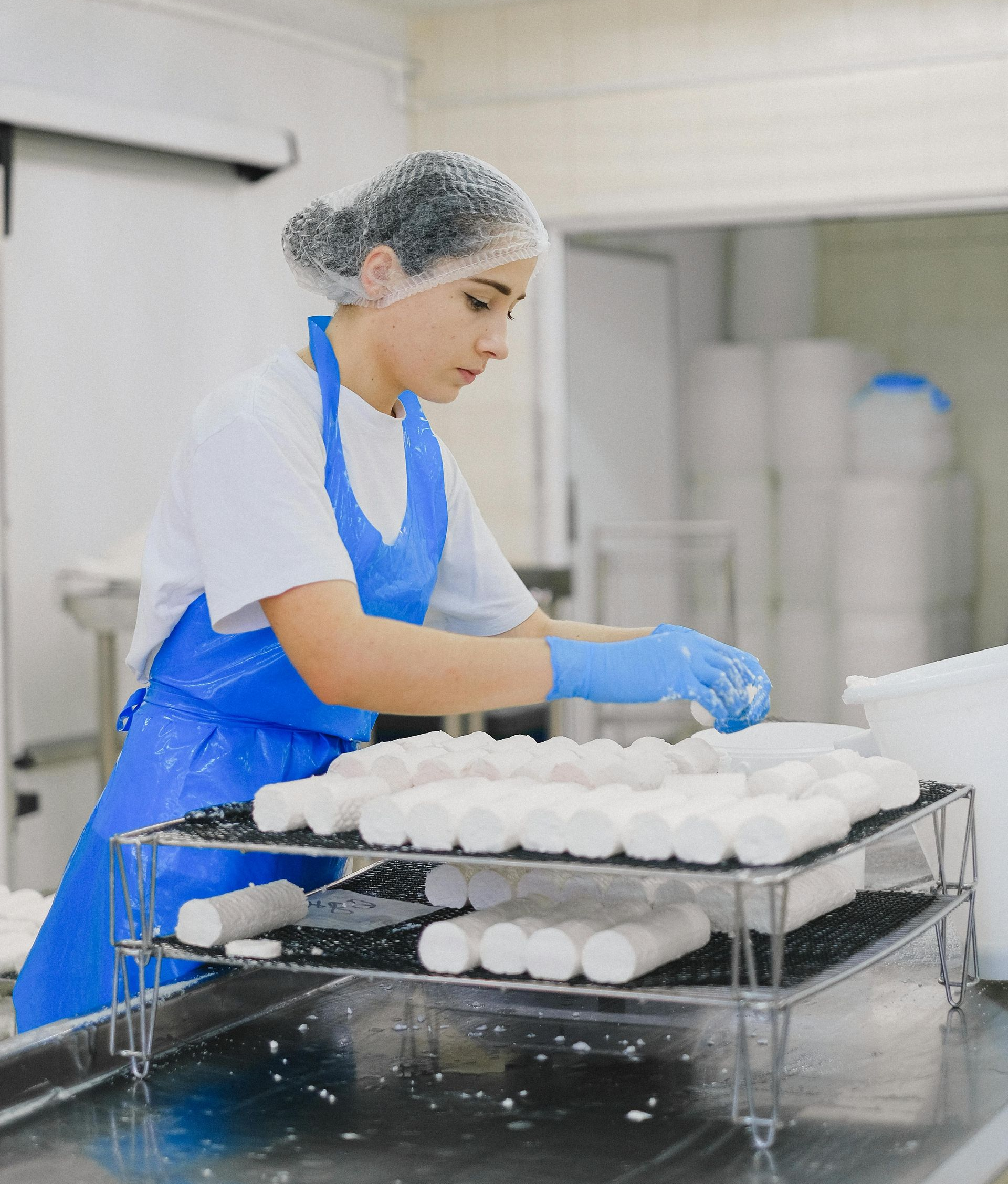 Woman in a clean room arranging white cheese rolls, wearing apron, hairnet, and gloves.