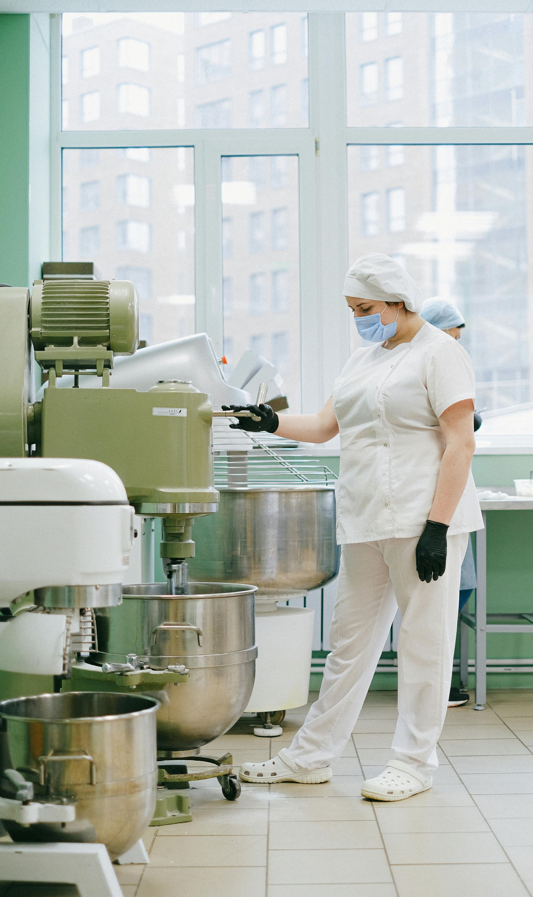 Woman in white uniform and mask operates industrial mixer in a bakery kitchen.