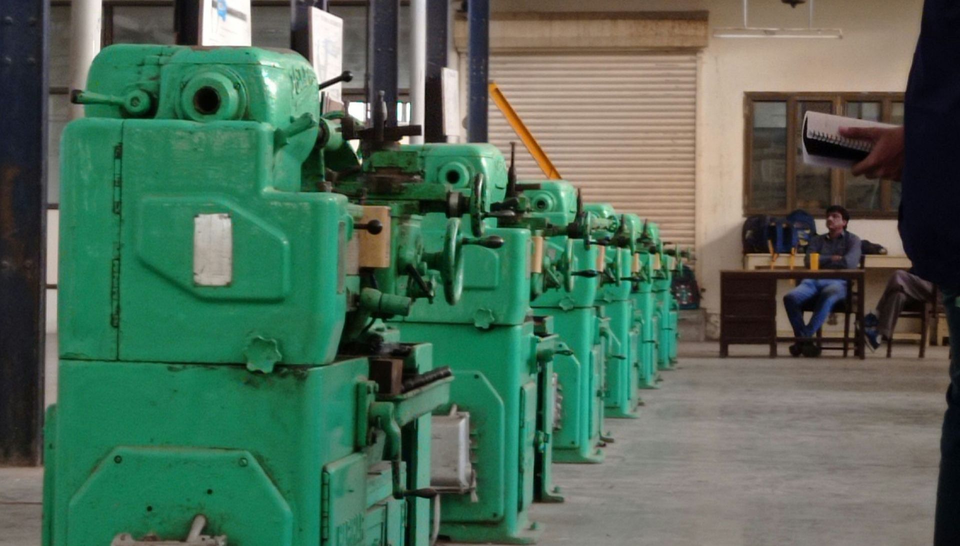 Row of green industrial machines in a workshop. A person holds papers in the foreground, another sits at a desk in the background.