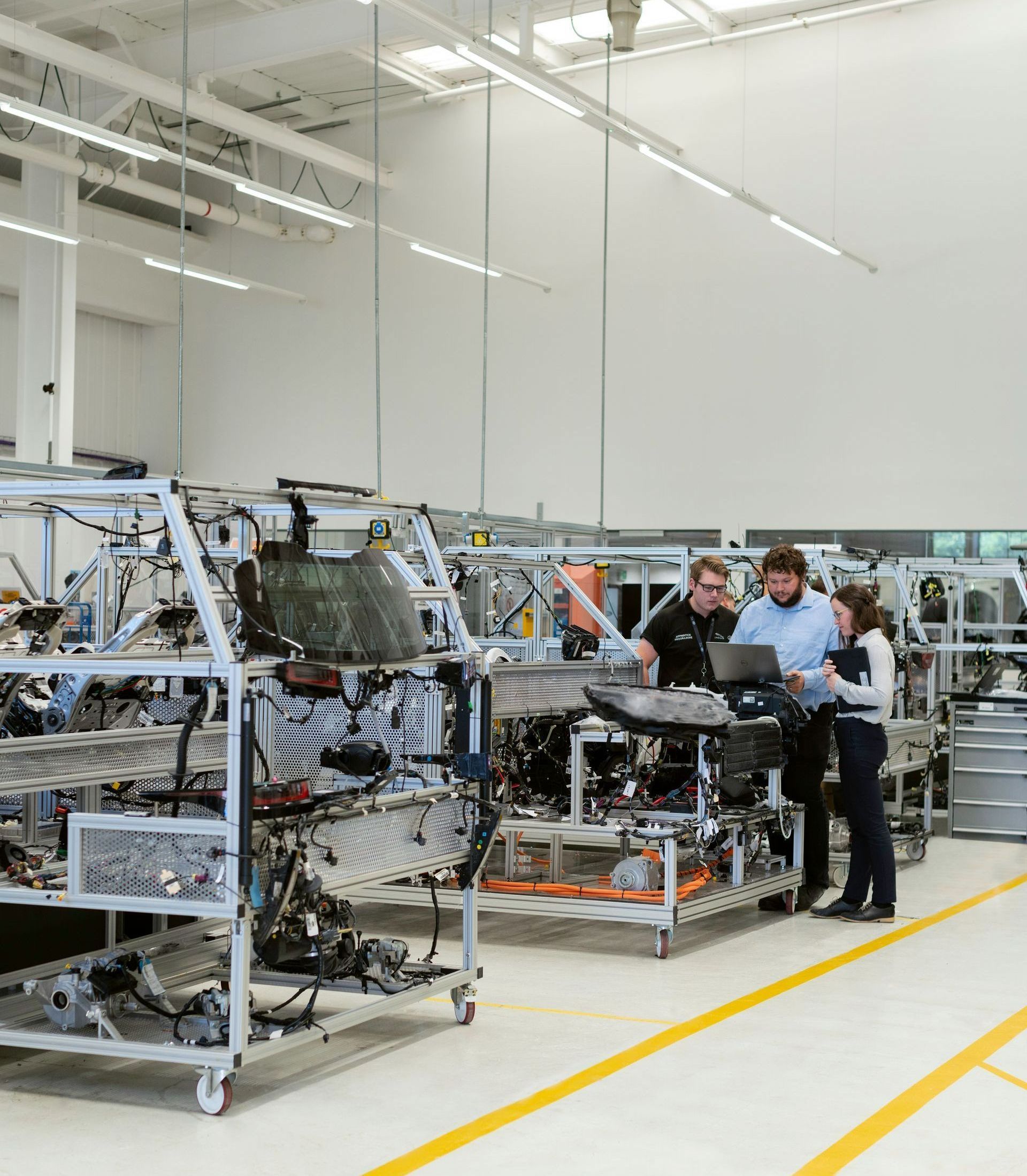 Workers reviewing a laptop next to car frames on a factory floor. Bright lighting and yellow lines highlight the setting.