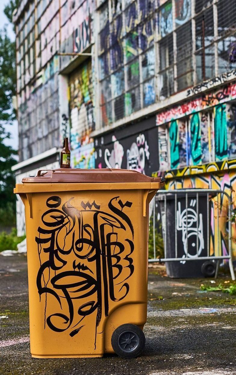 A yellow trash can with graffiti on it is sitting in front of a graffiti covered building.