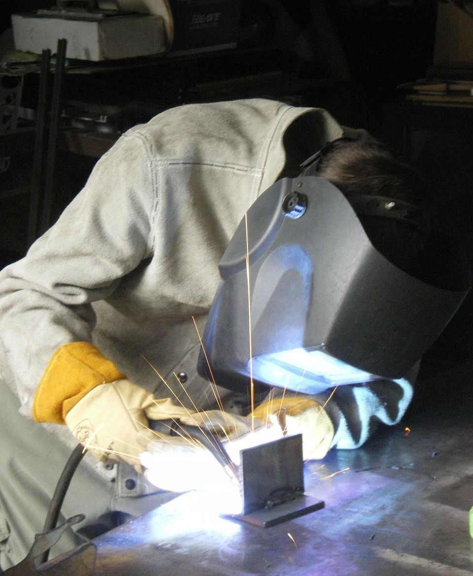 Welder in protective gear welding metal; sparks fly, creating a bright light.