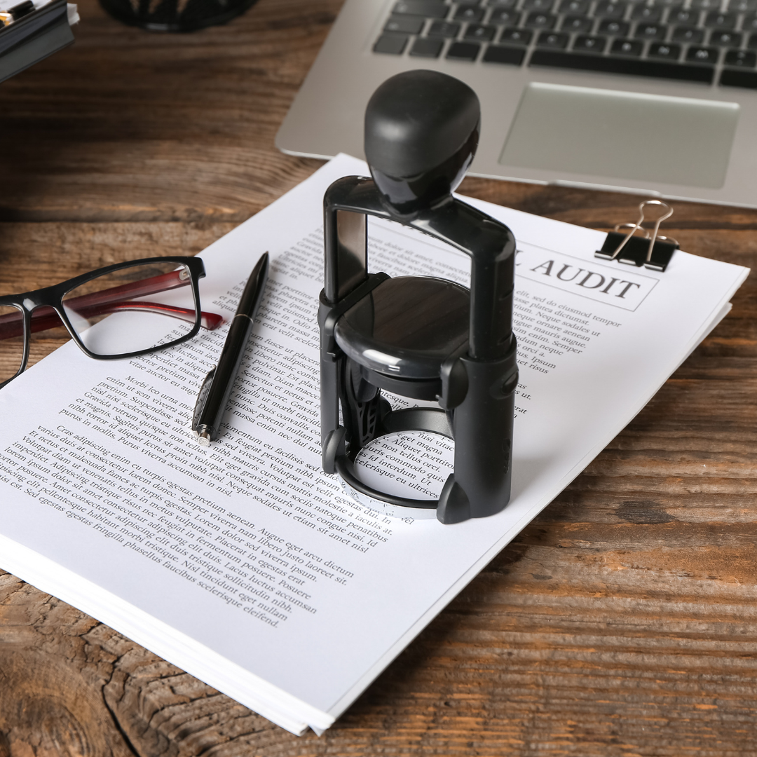 Black stamp over audit document on wooden desk, next to glasses, pen, and laptop.