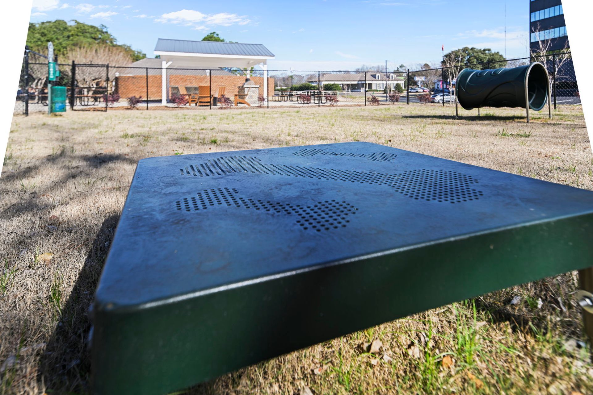 Low-angle view of a dark green pet play platform in a grassy dog park with a tunnel and pavilion in the background.