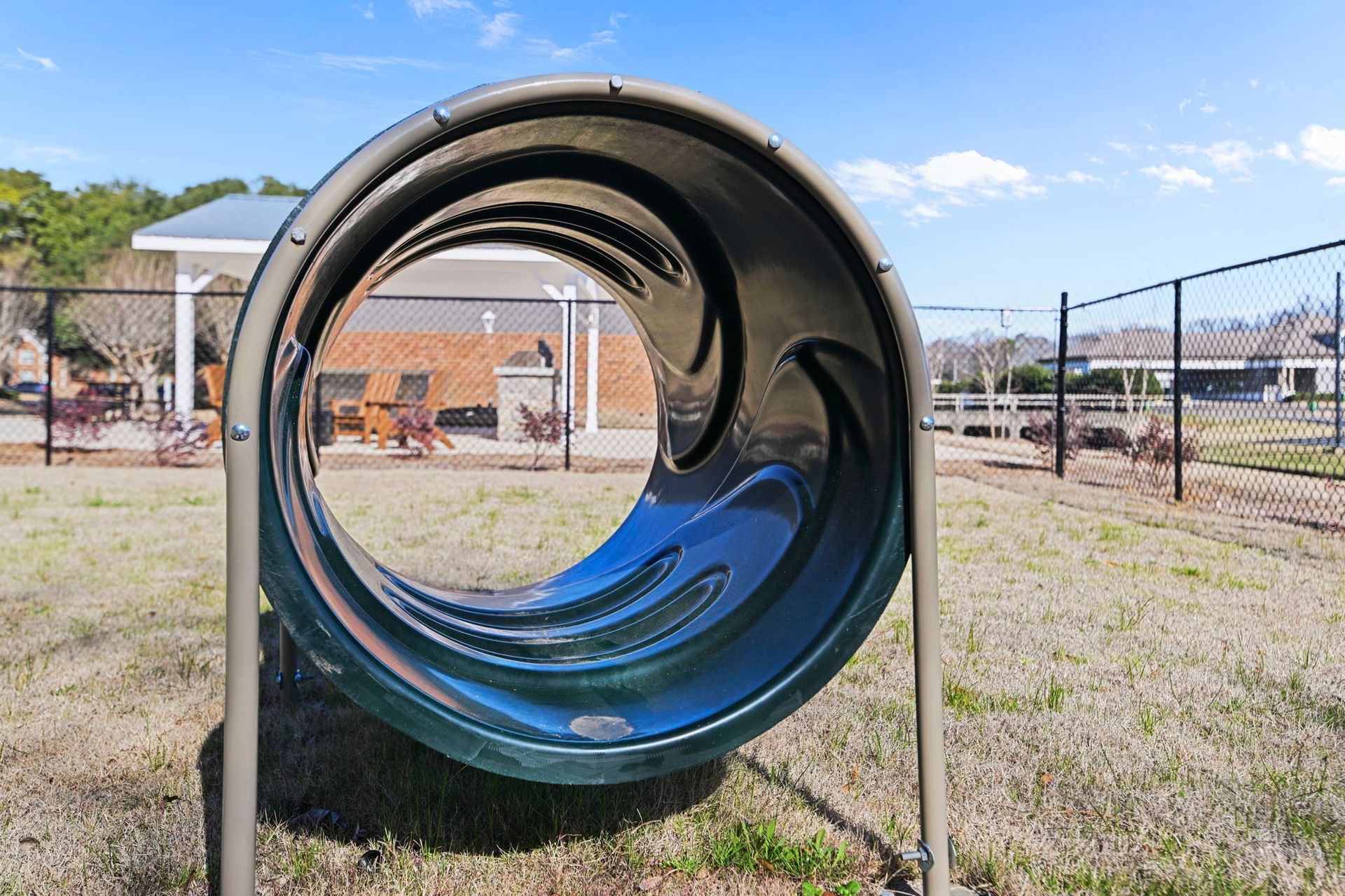 A dark green, cylindrical dog agility tunnel standing on a lawn in an outdoor park area under a blue sky.