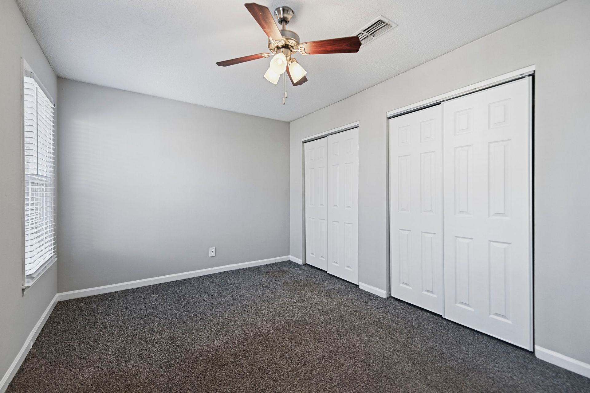 An empty bedroom with gray walls, gray carpet, a ceiling fan, a window with blinds, and two sets of white closet doors.