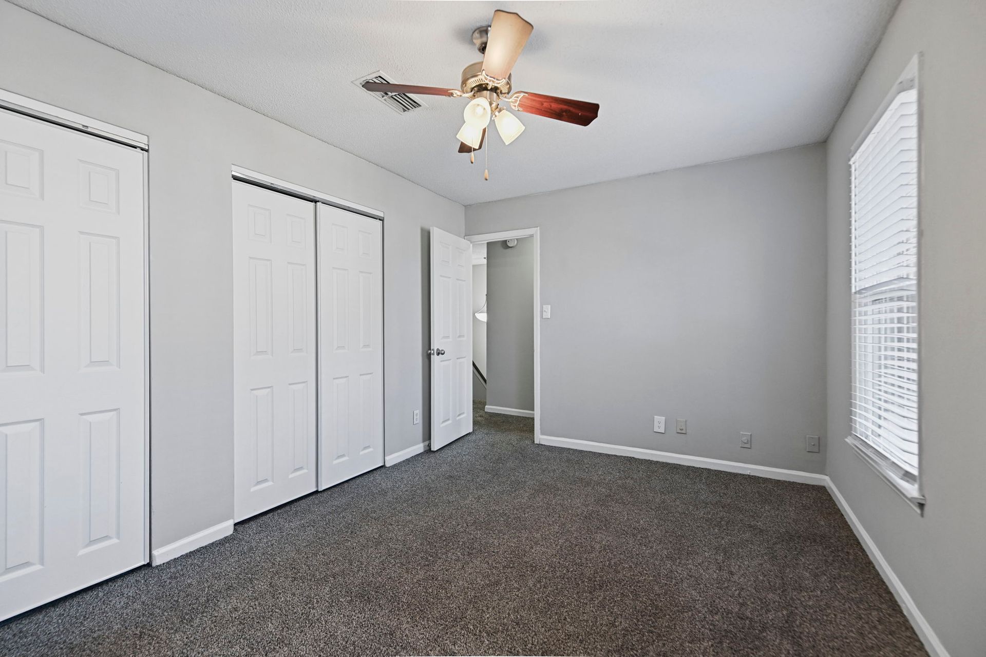 An empty bedroom with light gray walls, dark carpet, a white ceiling fan, and white closet doors with a window.