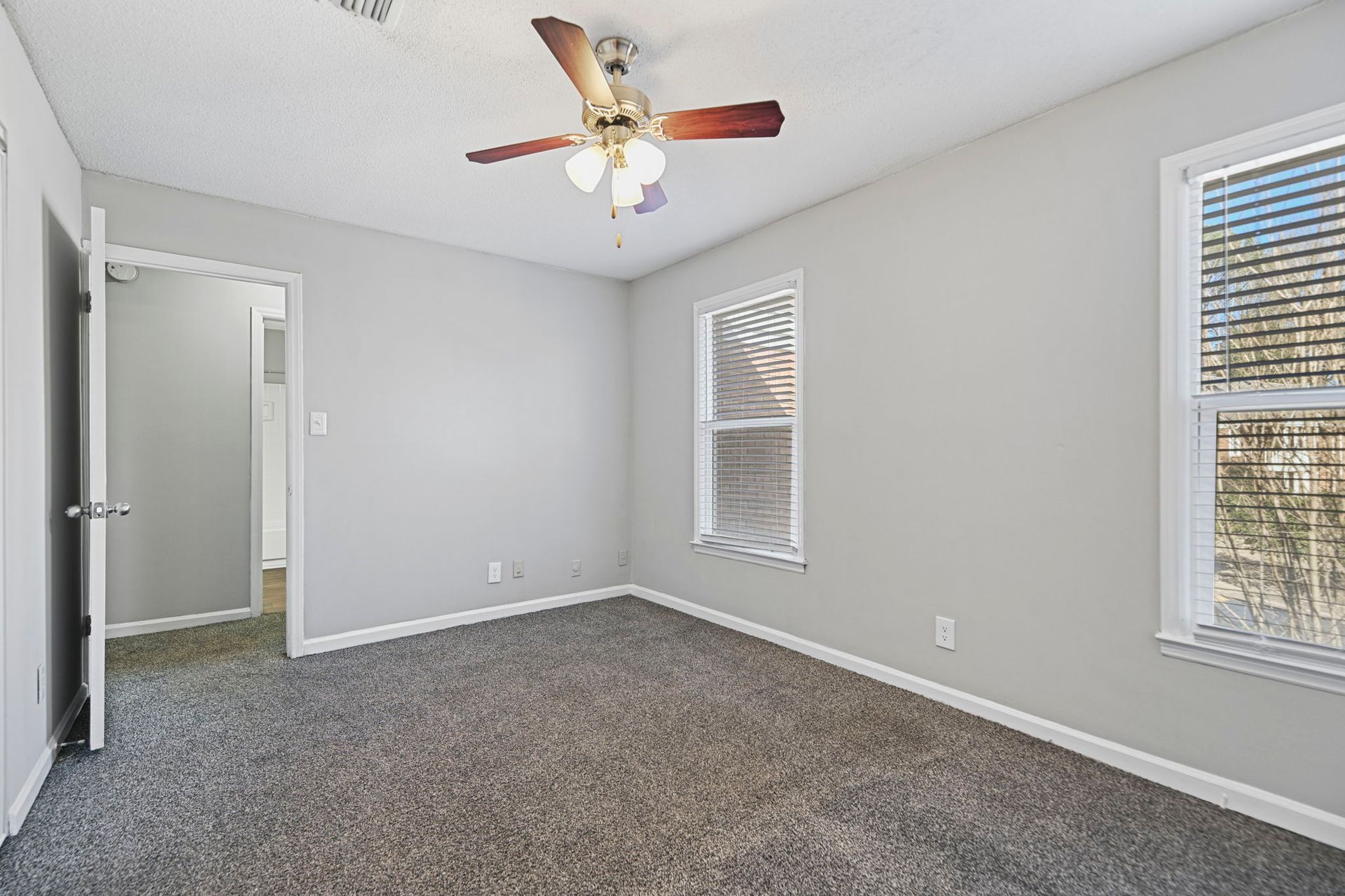 Empty bedroom with light gray walls, gray carpet, a ceiling fan, and two windows with blinds.