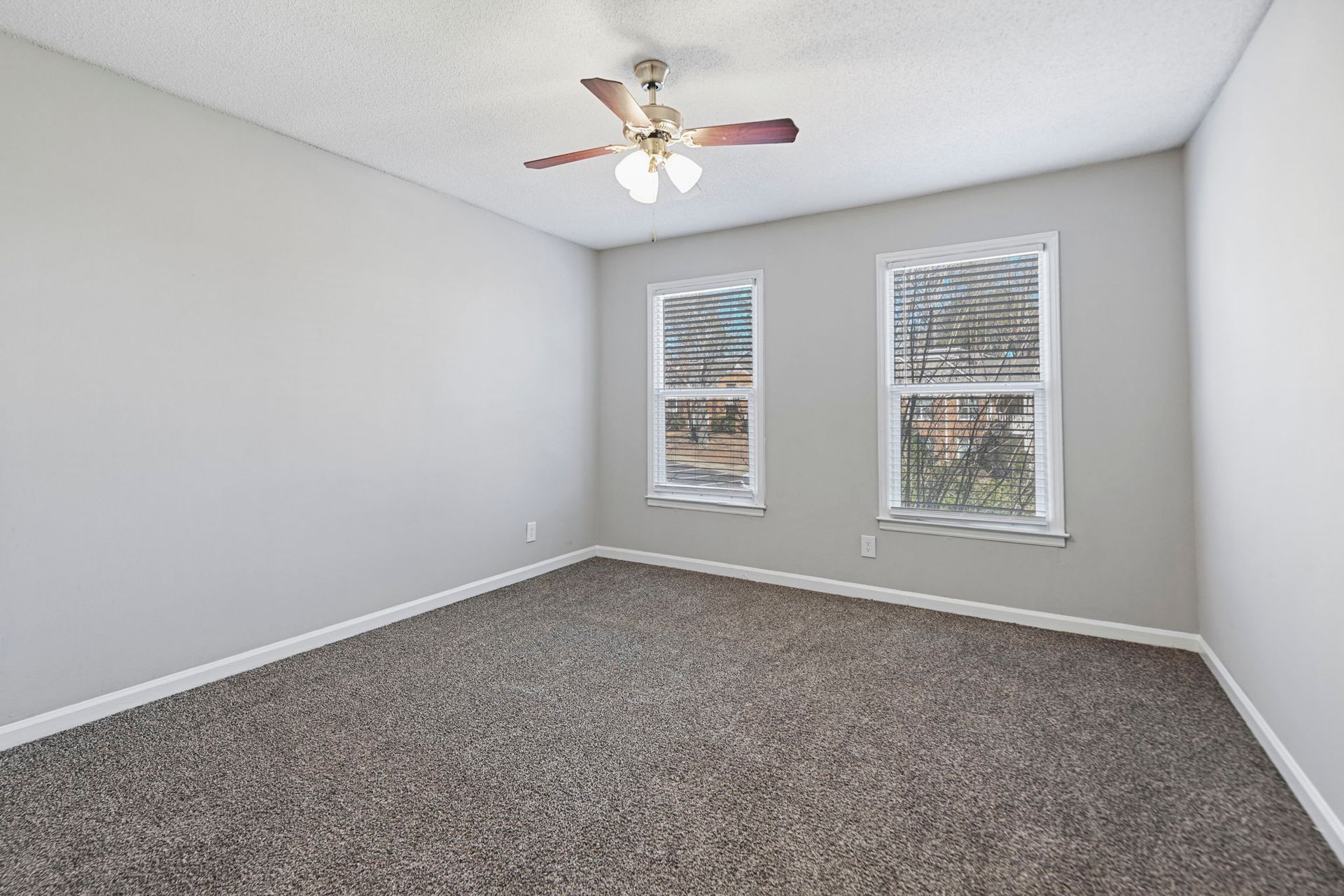 An empty room with light gray walls, brown carpet, a ceiling fan, and two windows looking out onto trees.
