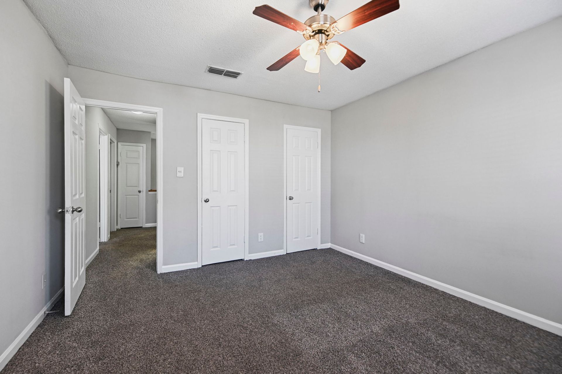 A light gray bedroom with gray carpet, a ceiling fan, and two white closed doors leading to a hallway.