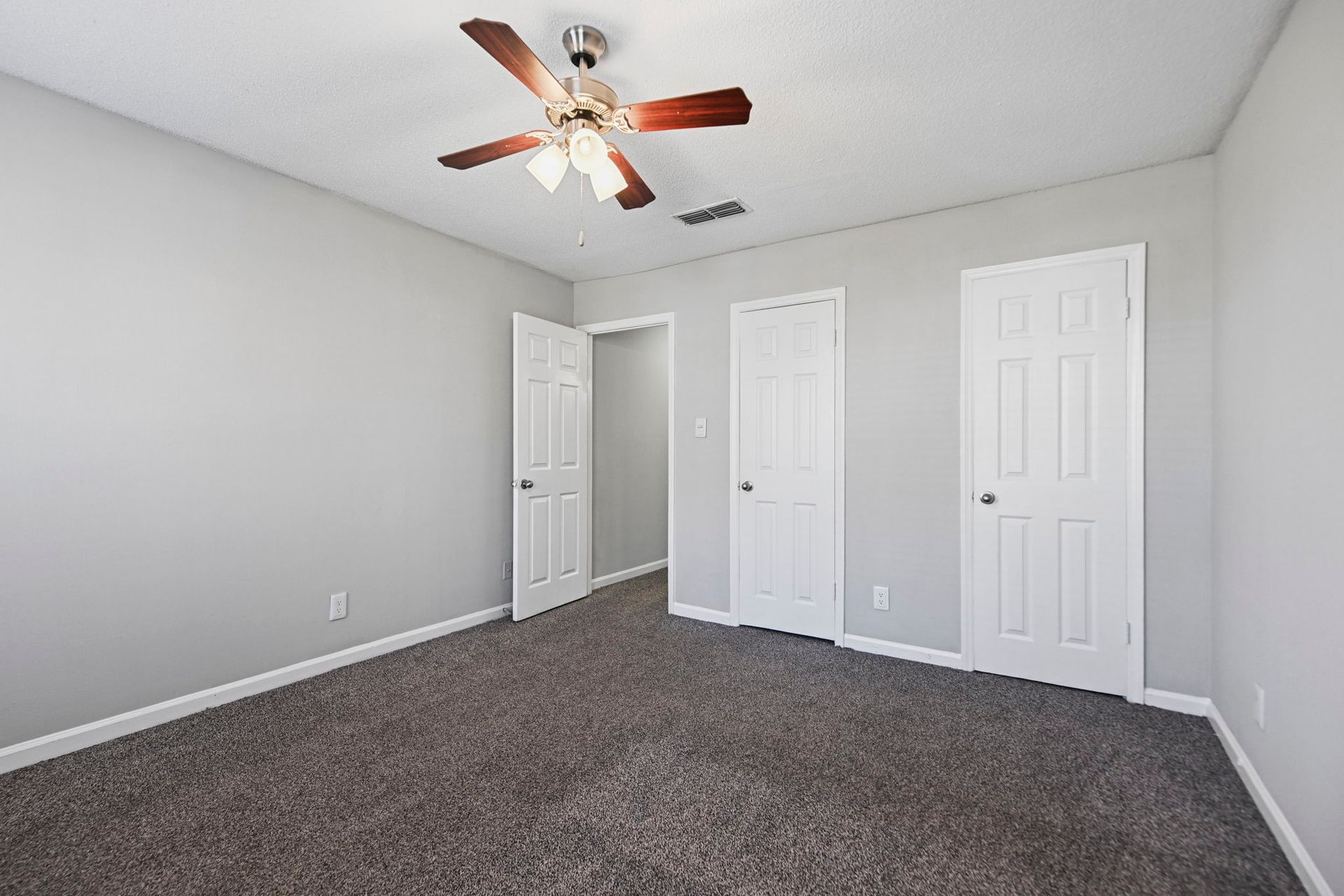 An empty room with light gray walls, dark gray carpet, a ceiling fan, and three white doors leading to other areas.