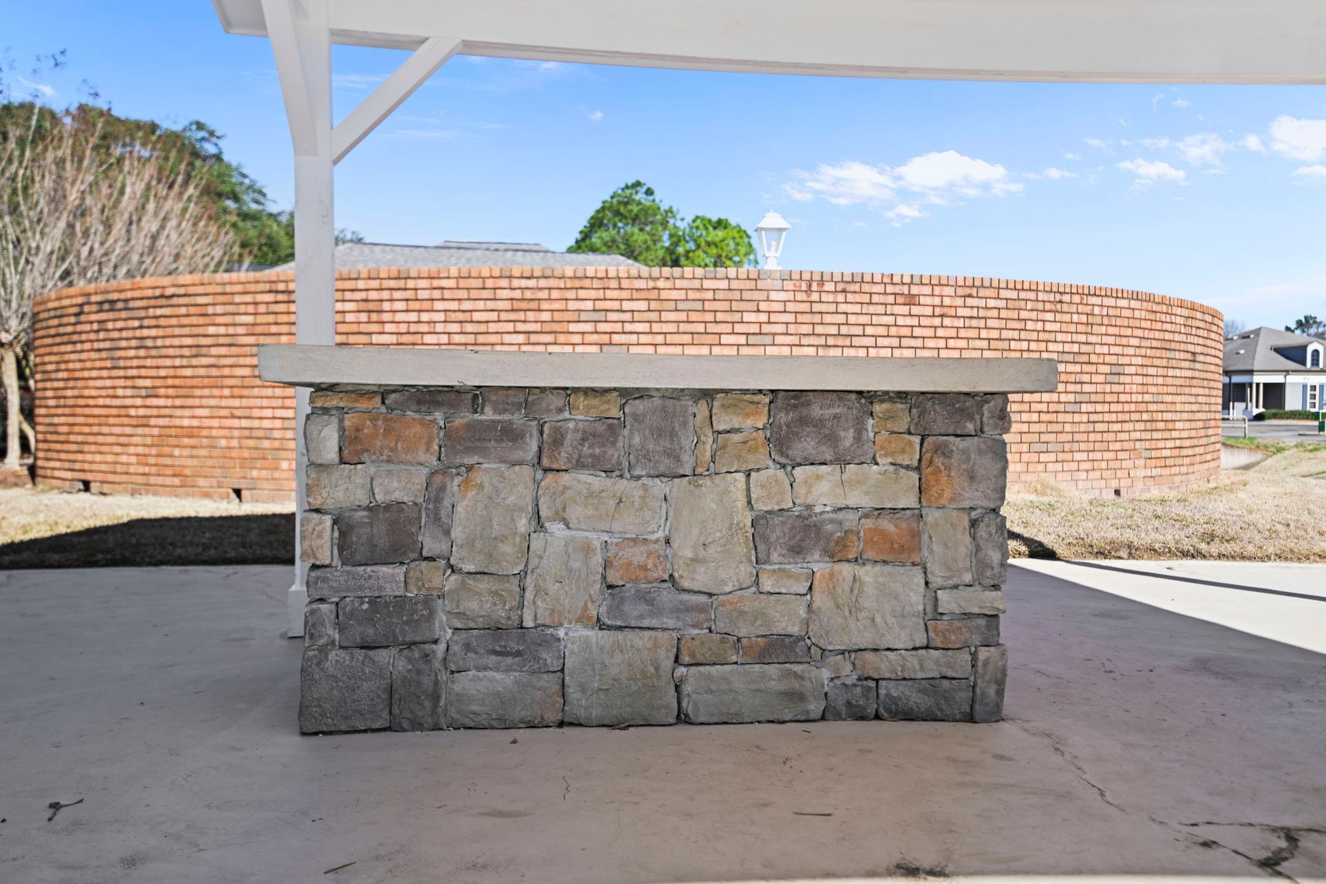 A stone-faced outdoor bar sits on a concrete patio under a white pavilion, with a large, curved brick wall in the distance.