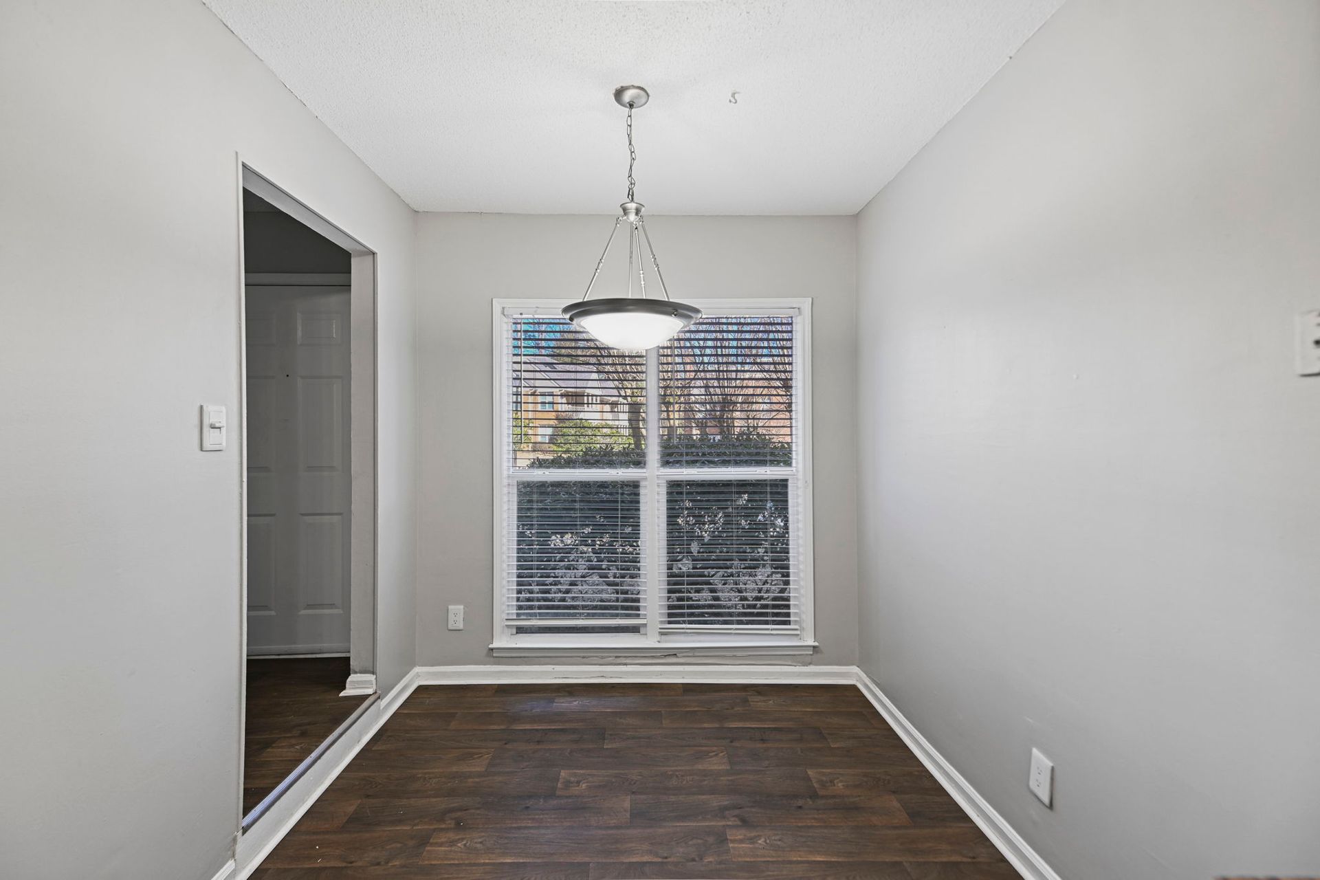 A small, neutral-toned dining nook with dark wood flooring, a central pendant light, and a window with plantation shutters.