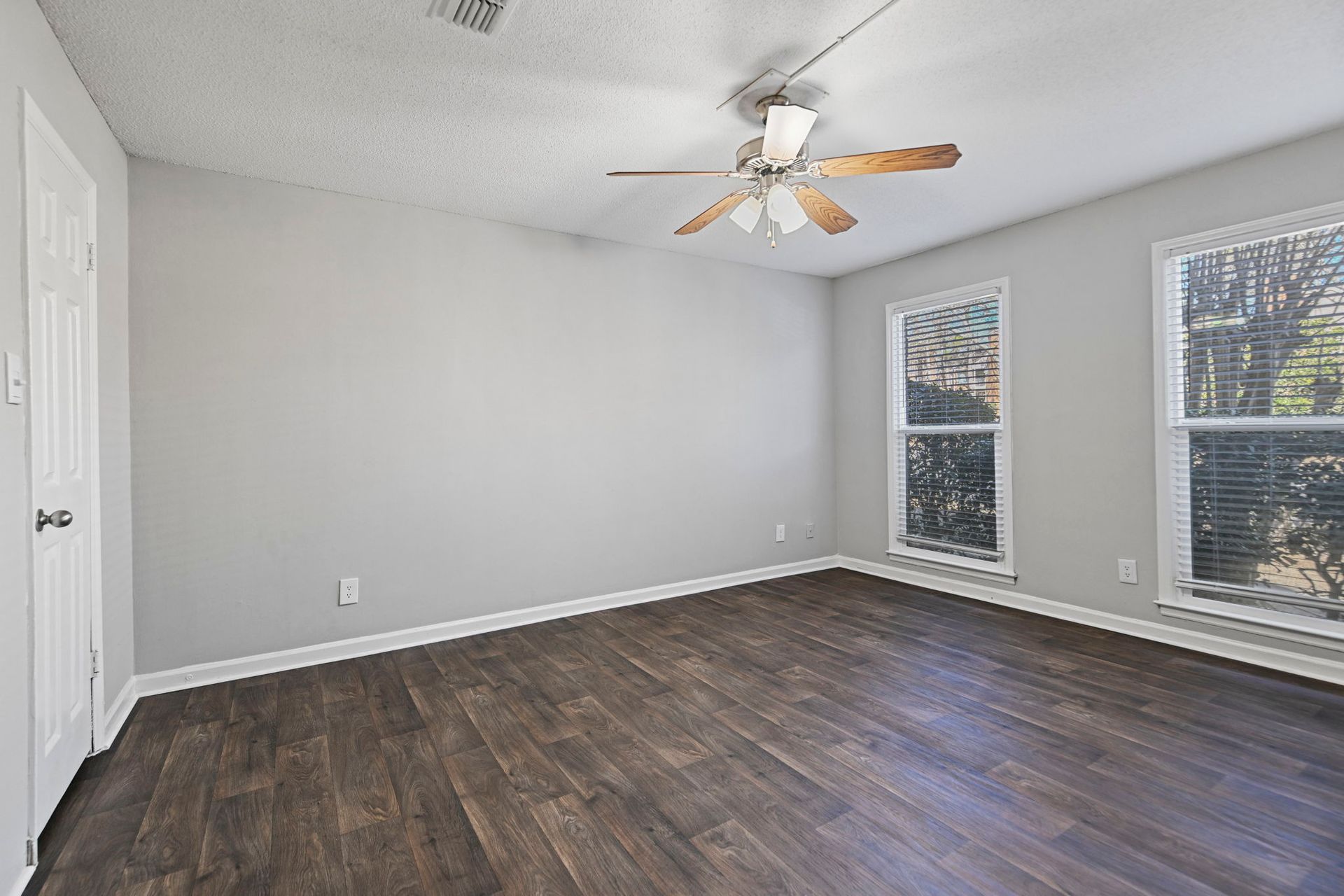 Empty bedroom with light grey walls, a ceiling fan, dark wood floors, and two large windows.