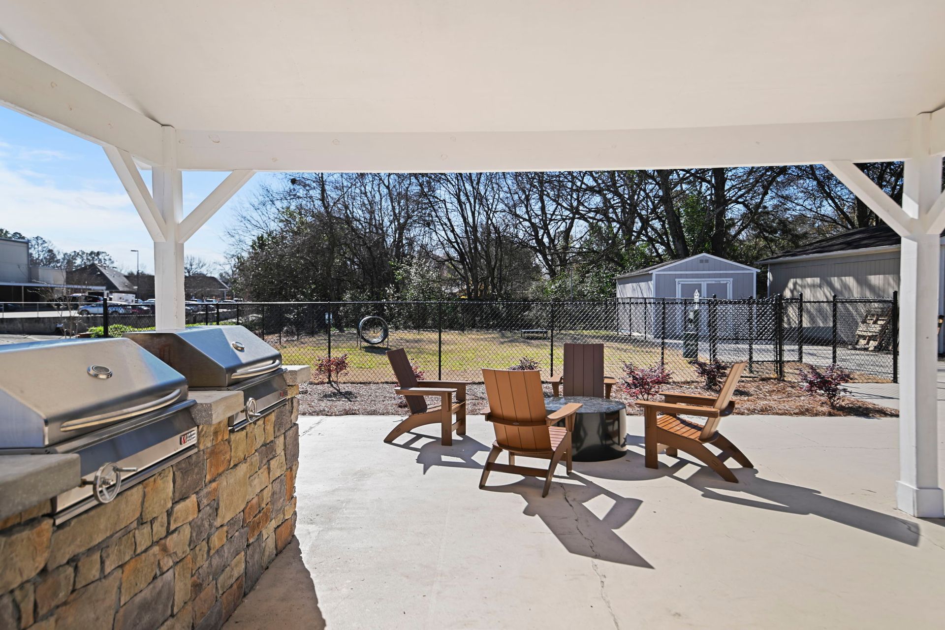 Covered patio with a stone grill station, three wooden chairs, and a fire pit, overlooking a fenced backyard.