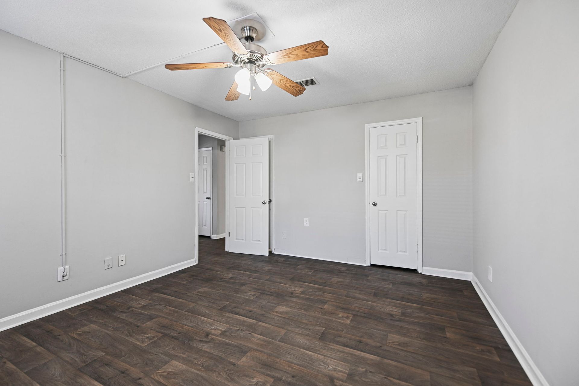 An empty bedroom with gray walls, dark wood-look flooring, two white doors, and a ceiling fan with lights.