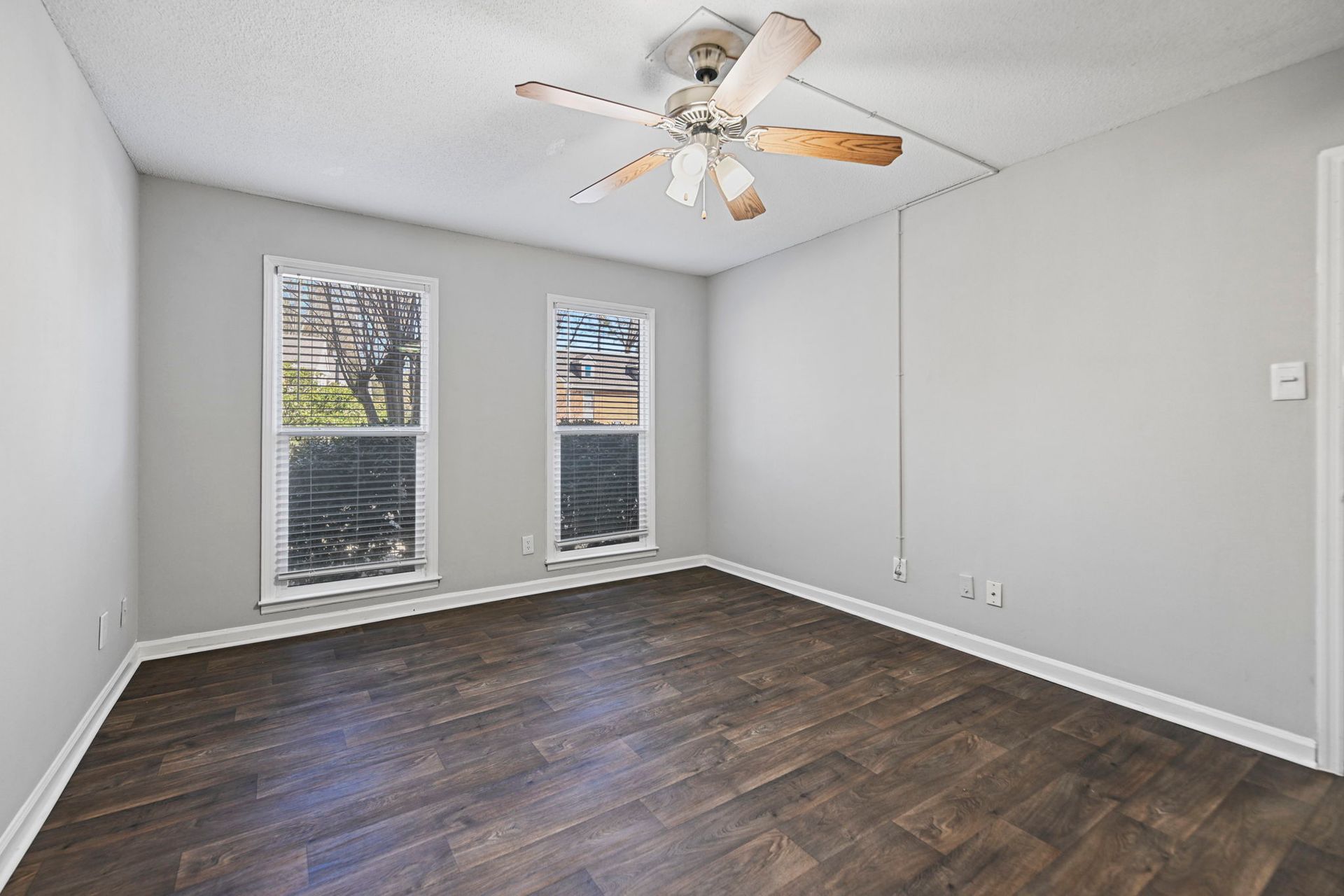 An empty room with light gray walls, dark wood-look flooring, two windows, and a white ceiling fan.