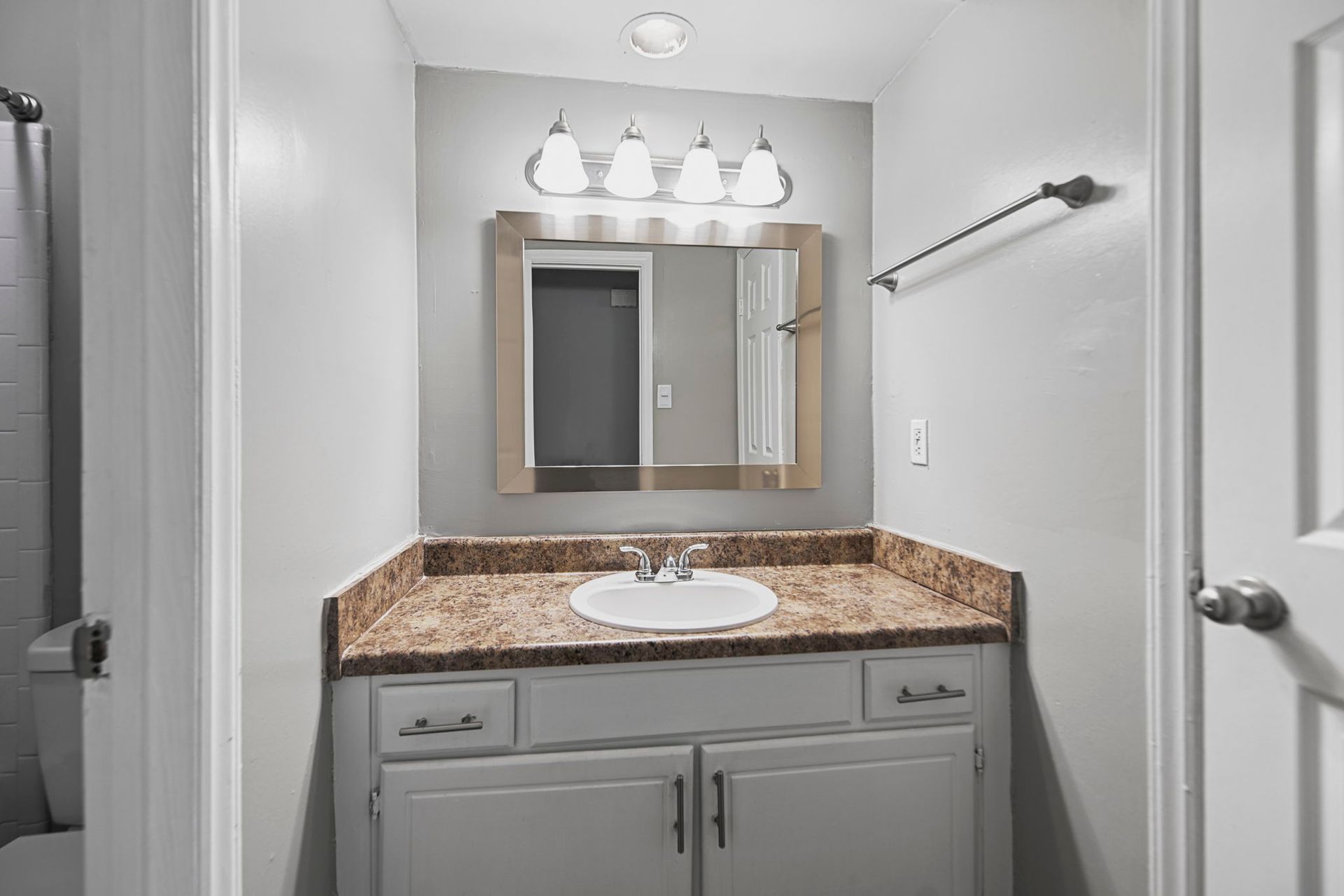 A bathroom vanity with a speckled brown countertop, light gray cabinets, a rectangular mirror, and overhead lighting.