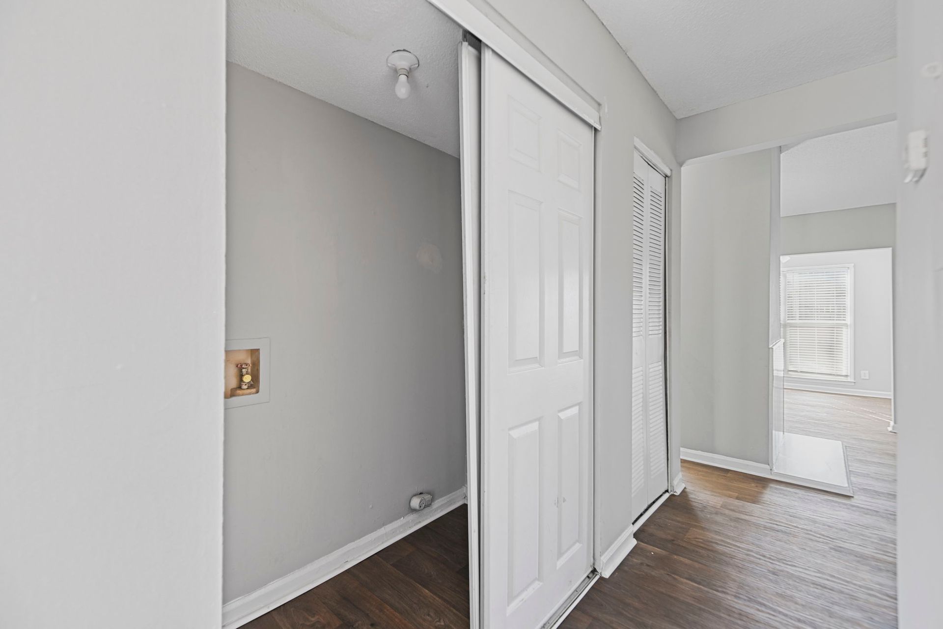 A hallway with light gray walls, dark wood-look flooring, and a white sliding door leading to an adjacent room.