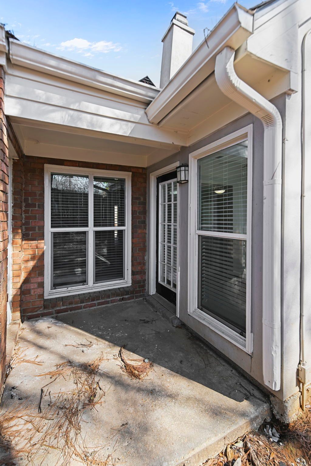 A gray-toned home exterior featuring a small concrete patio, a brick wall, two windows with blinds, and a glass door.