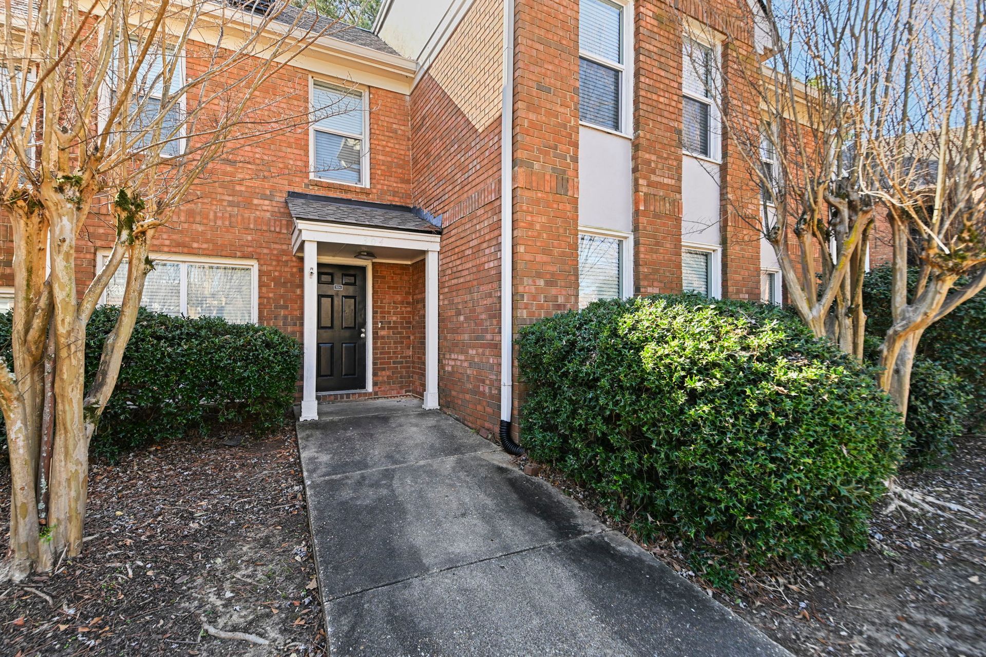 A brick multi-unit building with a black front door, a concrete walkway, and surrounding green bushes.