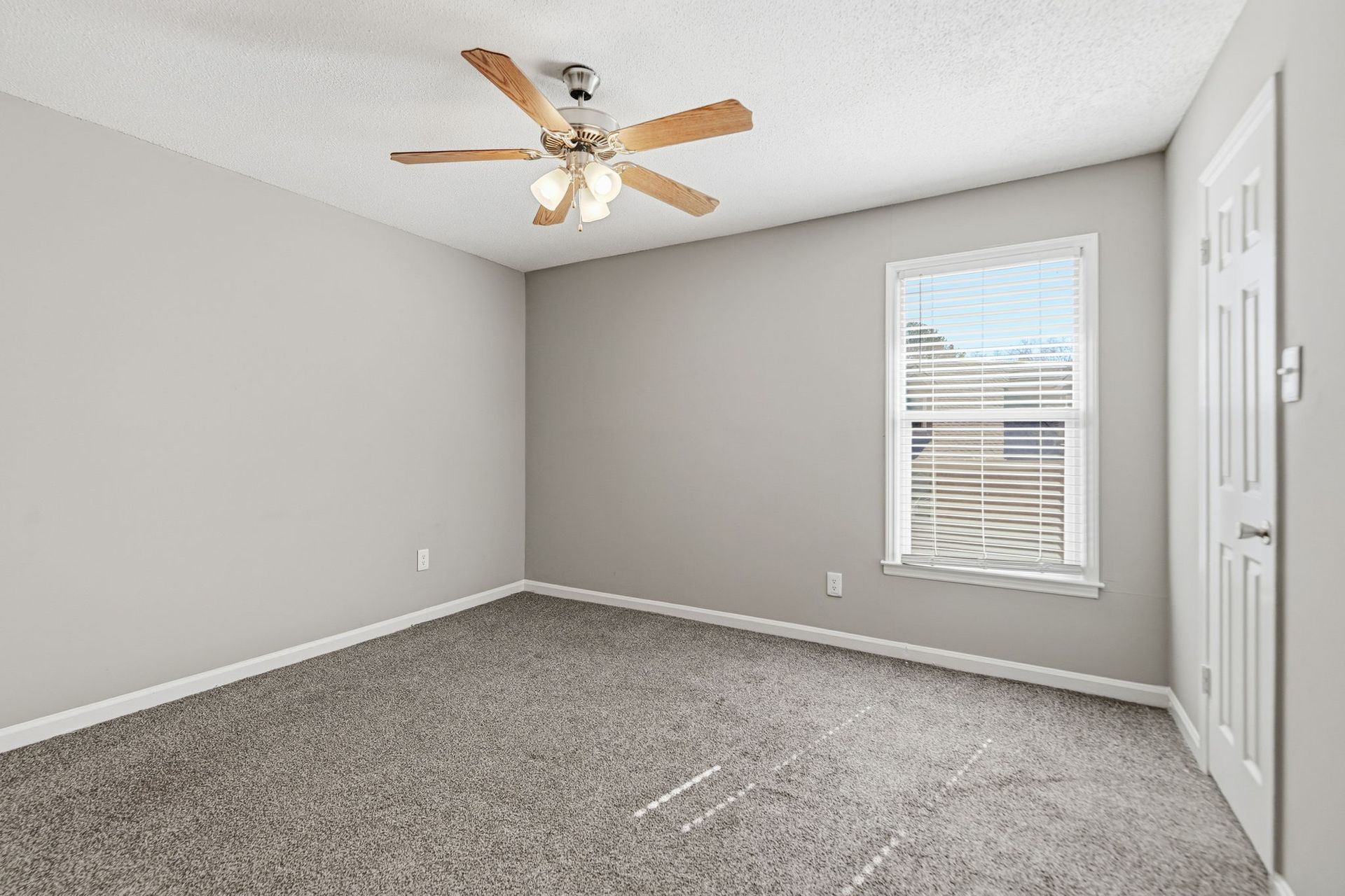 A neutral-colored bedroom featuring a ceiling fan, a single window with blinds, and light-colored carpeting.