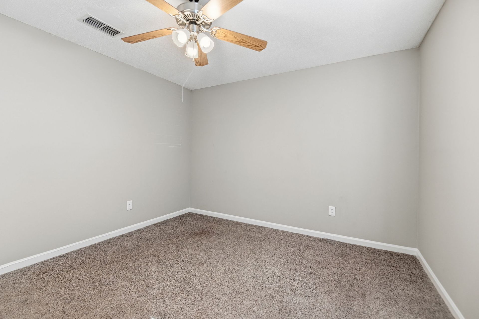 An empty room with light gray walls, beige carpet, and a wooden ceiling fan with a light fixture.