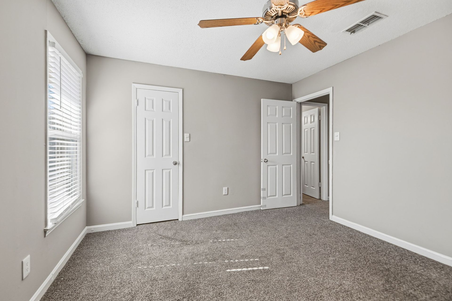 An empty bedroom with gray walls, beige carpet, a ceiling fan, a window with blinds, and two white doors.