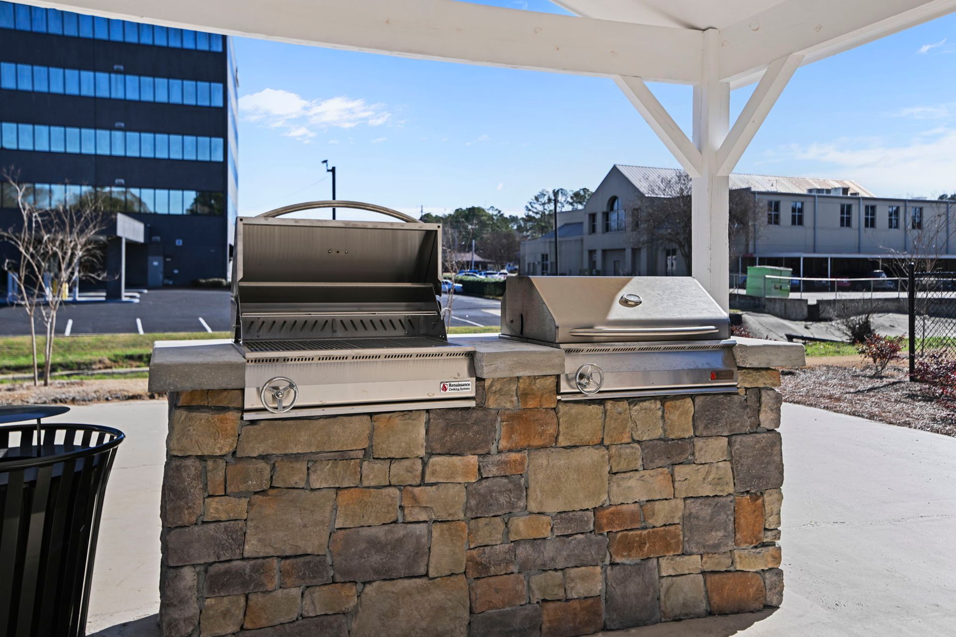 A stone outdoor grill station with two stainless steel grills under a white pergola, set against a sunny building exterior.