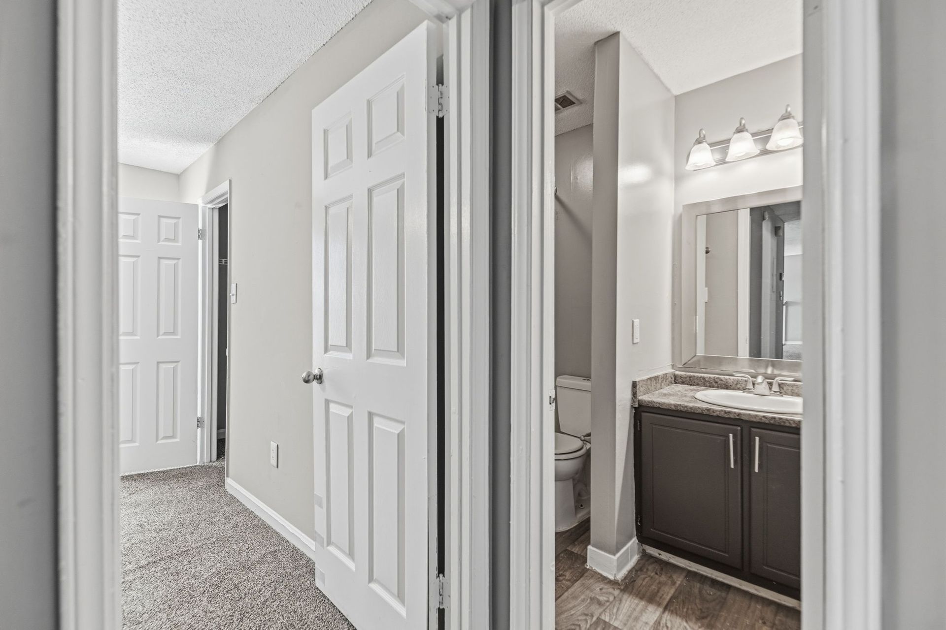 A view of a carpeted hallway leading to a door, next to an open doorway showing a bathroom with dark cabinets and a mirror.