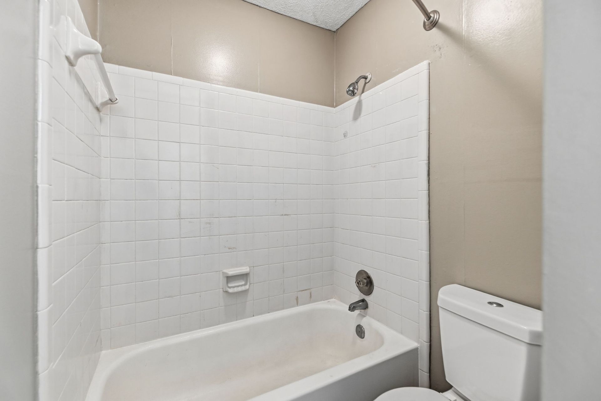 A white bathtub with white tiled walls and a showerhead, situated next to a white toilet in a bathroom with beige walls.