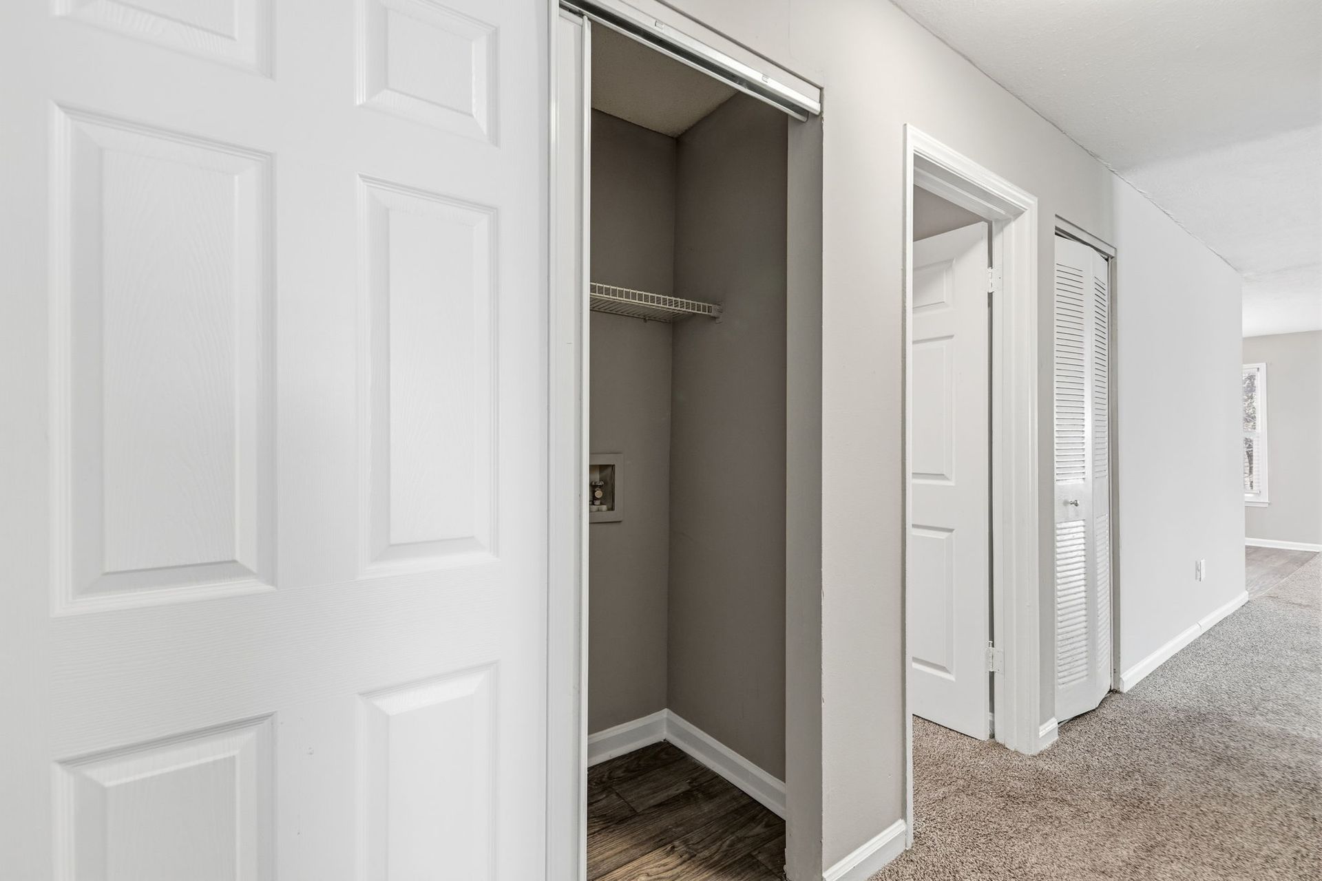 A hallway showing a small, open utility closet next to a closed white door and an open doorway leading to another room.