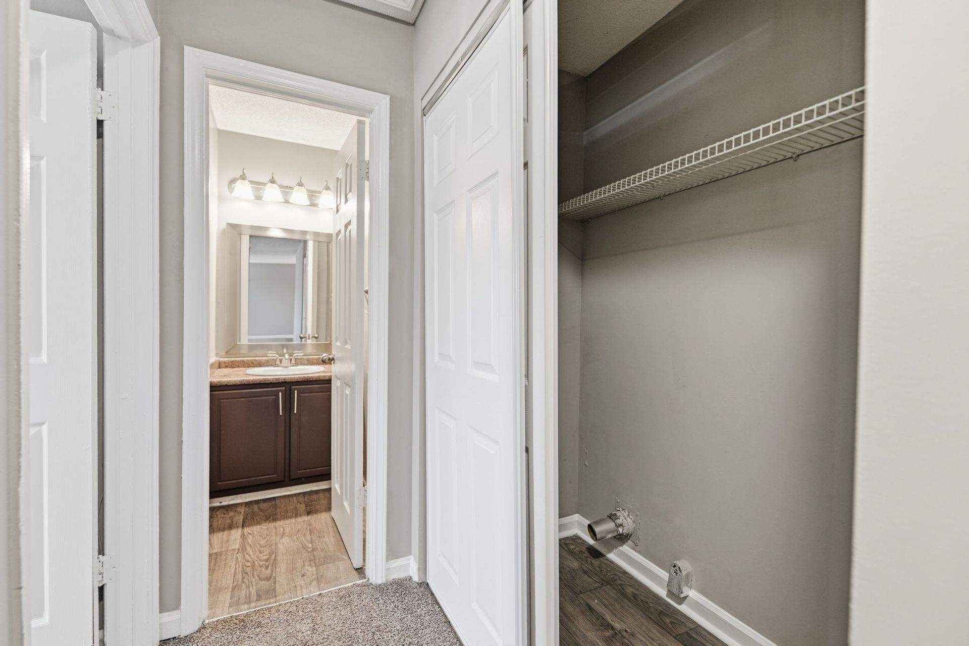 An open laundry closet with a shelf, next to a doorway showing a bathroom vanity with a mirror.
