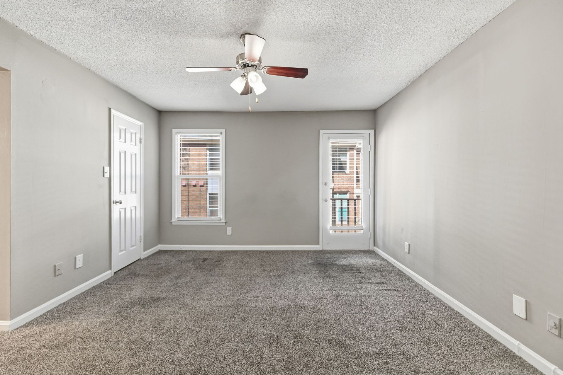 An empty room with light gray walls, textured carpet, a ceiling fan, a window, and a door leading to a small balcony.