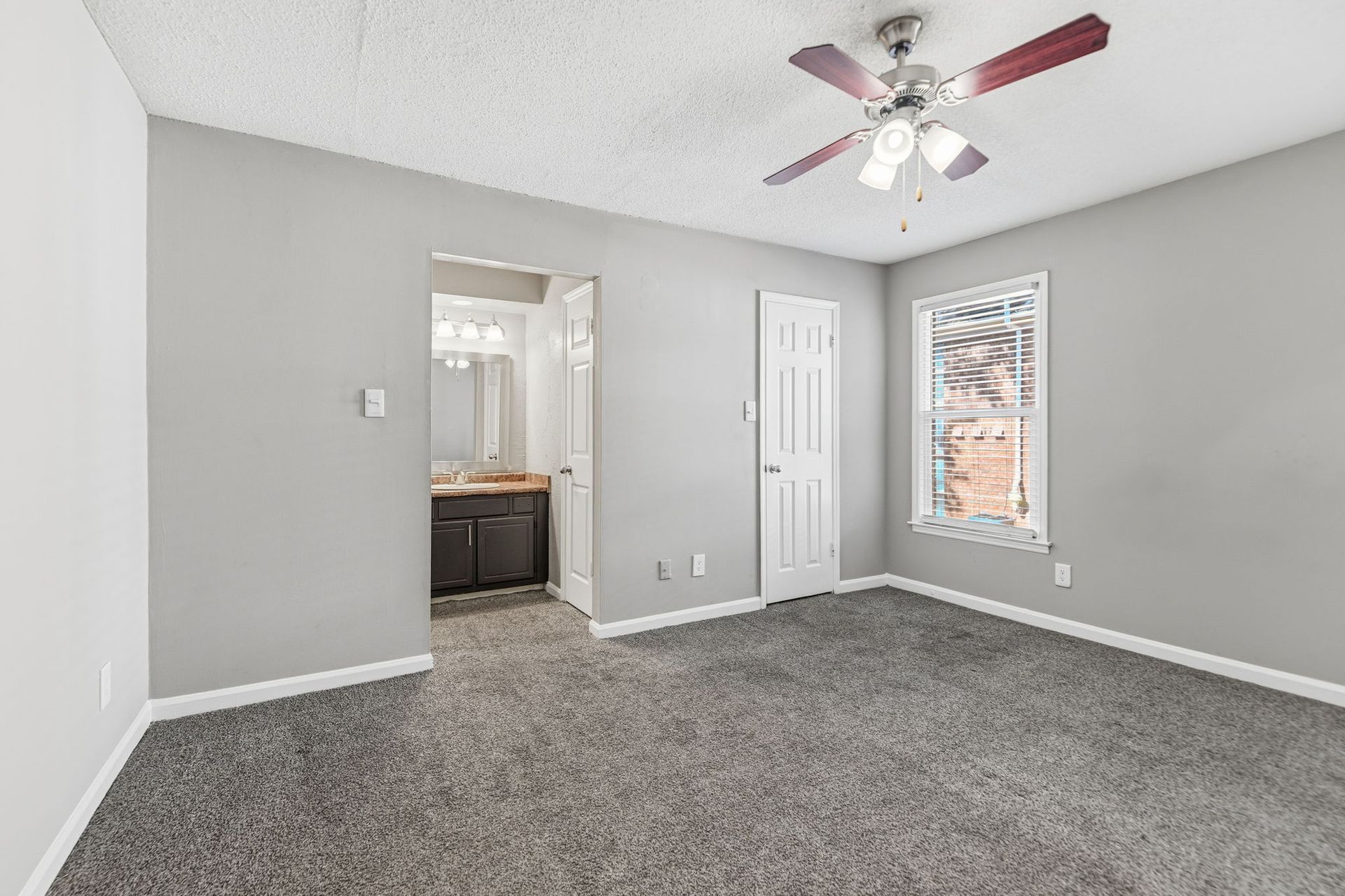 A vacant bedroom with gray walls, gray carpet, a white ceiling fan, a window, and an open doorway to a bathroom.