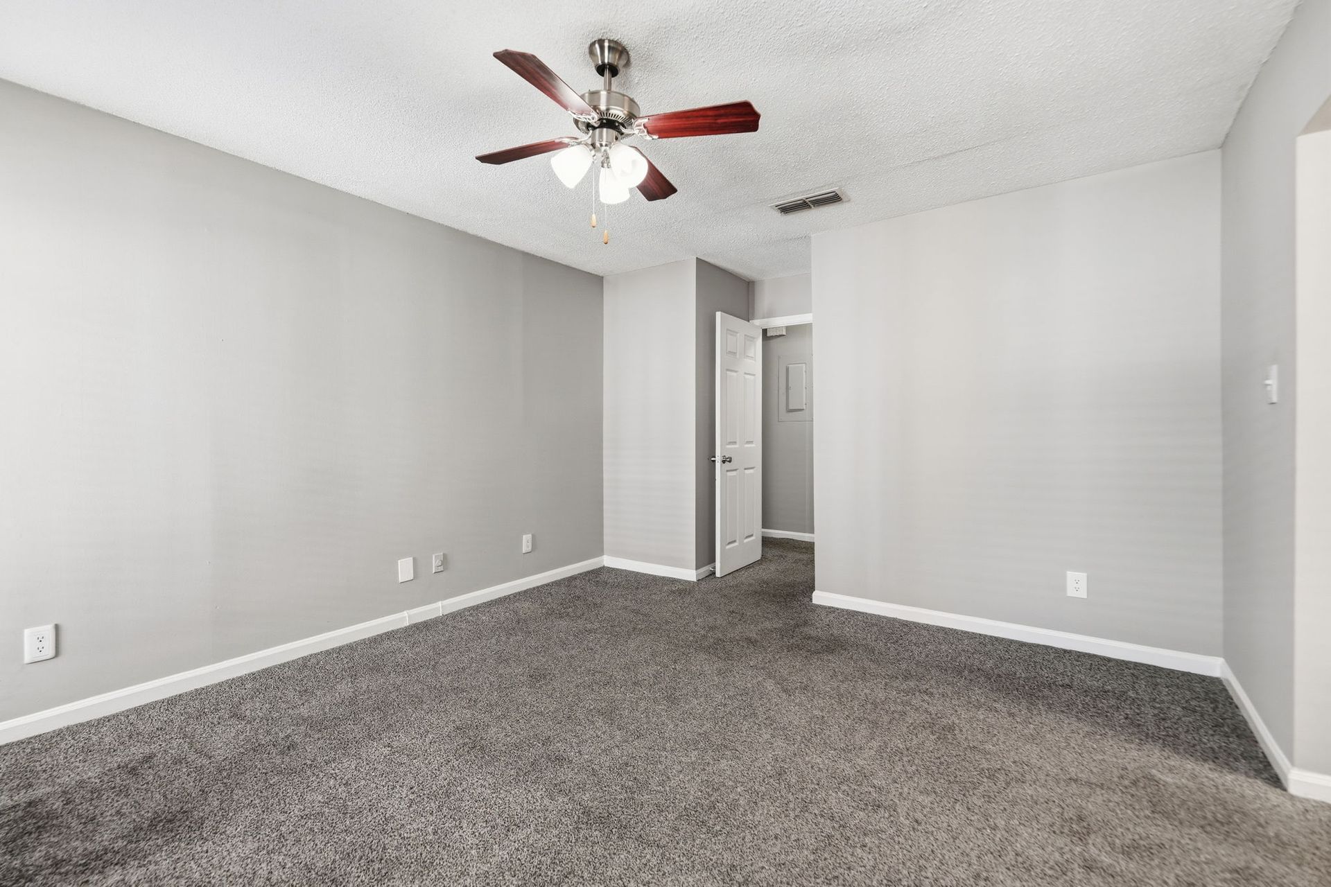 An empty room with gray walls, gray carpet, and a ceiling fan. A partially open white door leads to another room.