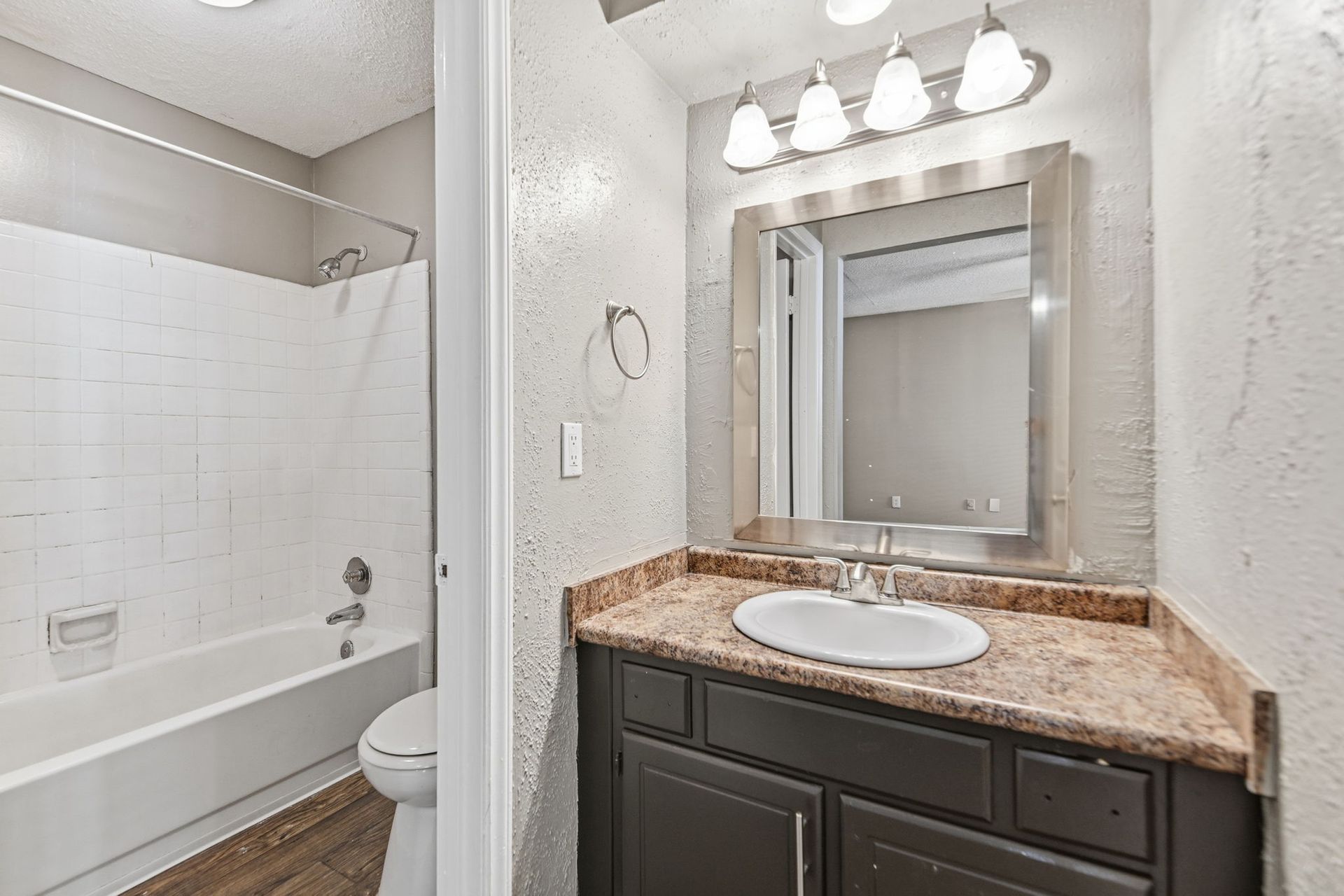 A bathroom featuring a white bathtub with a shower, a dark vanity with a tan countertop, and a mirror with light fixtures.