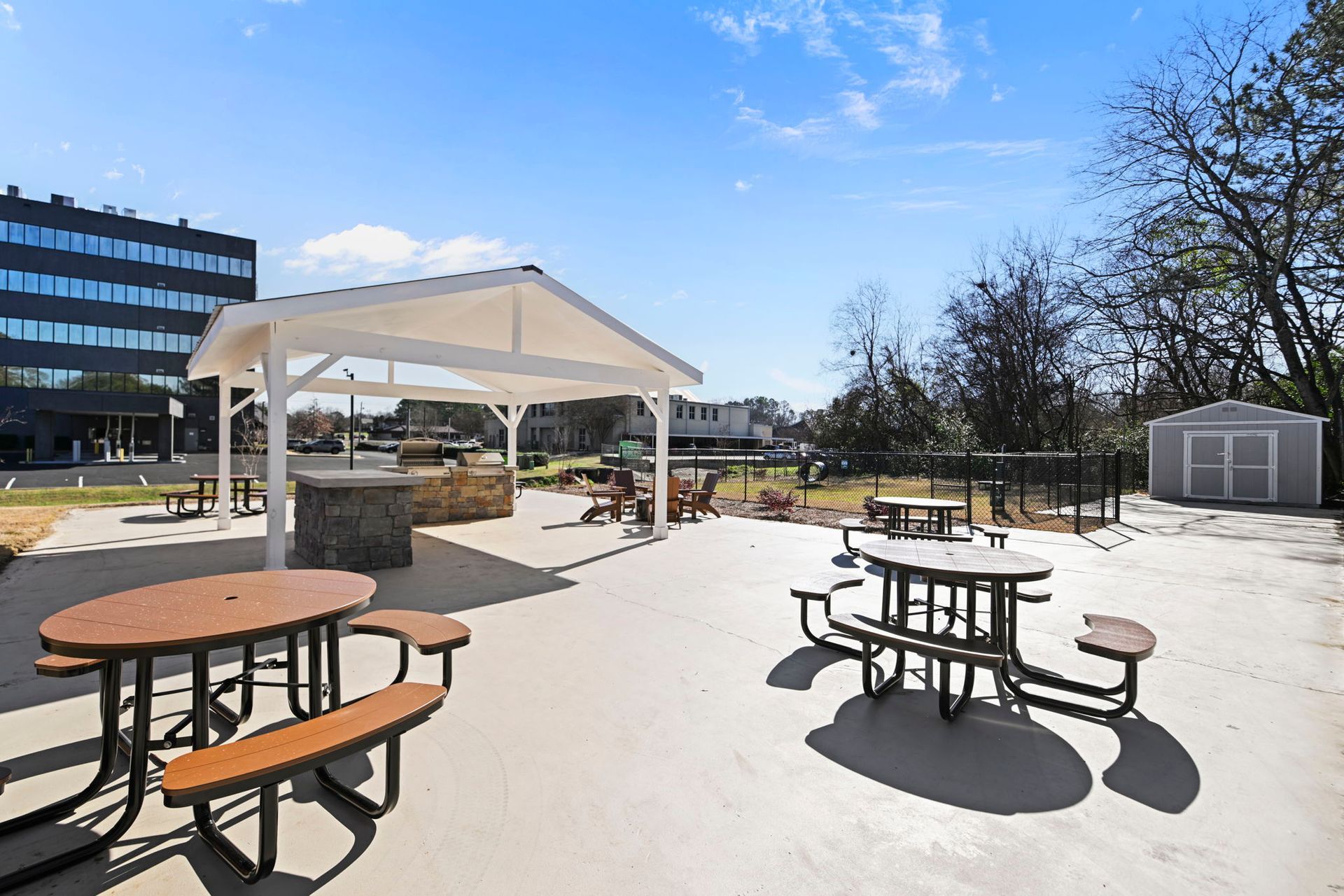 Outdoor patio area with a covered stone grilling station, multiple picnic tables, and a small storage shed under a blue sky.
